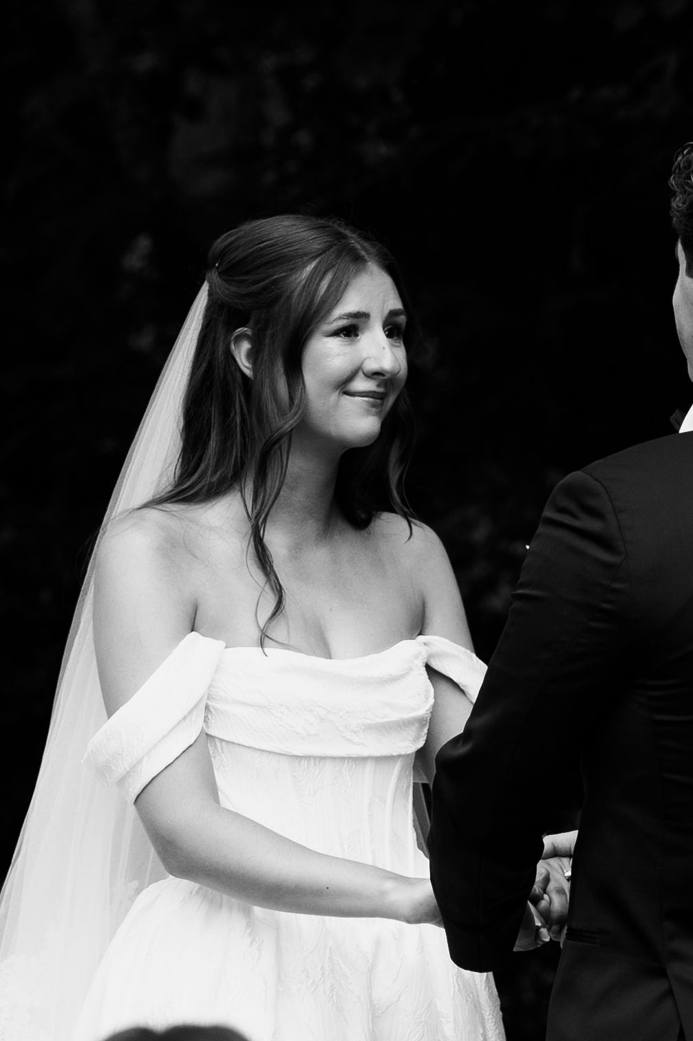 Black and white photo of a smiling bride in an off-shoulder wedding dress and veil holding hands at an outdoor ceremony.