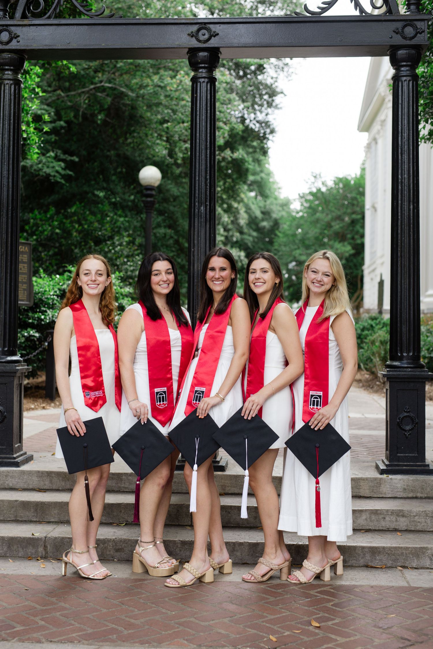 A group of graduates in white dresses and red sashes pose with diplomas under an archway.
