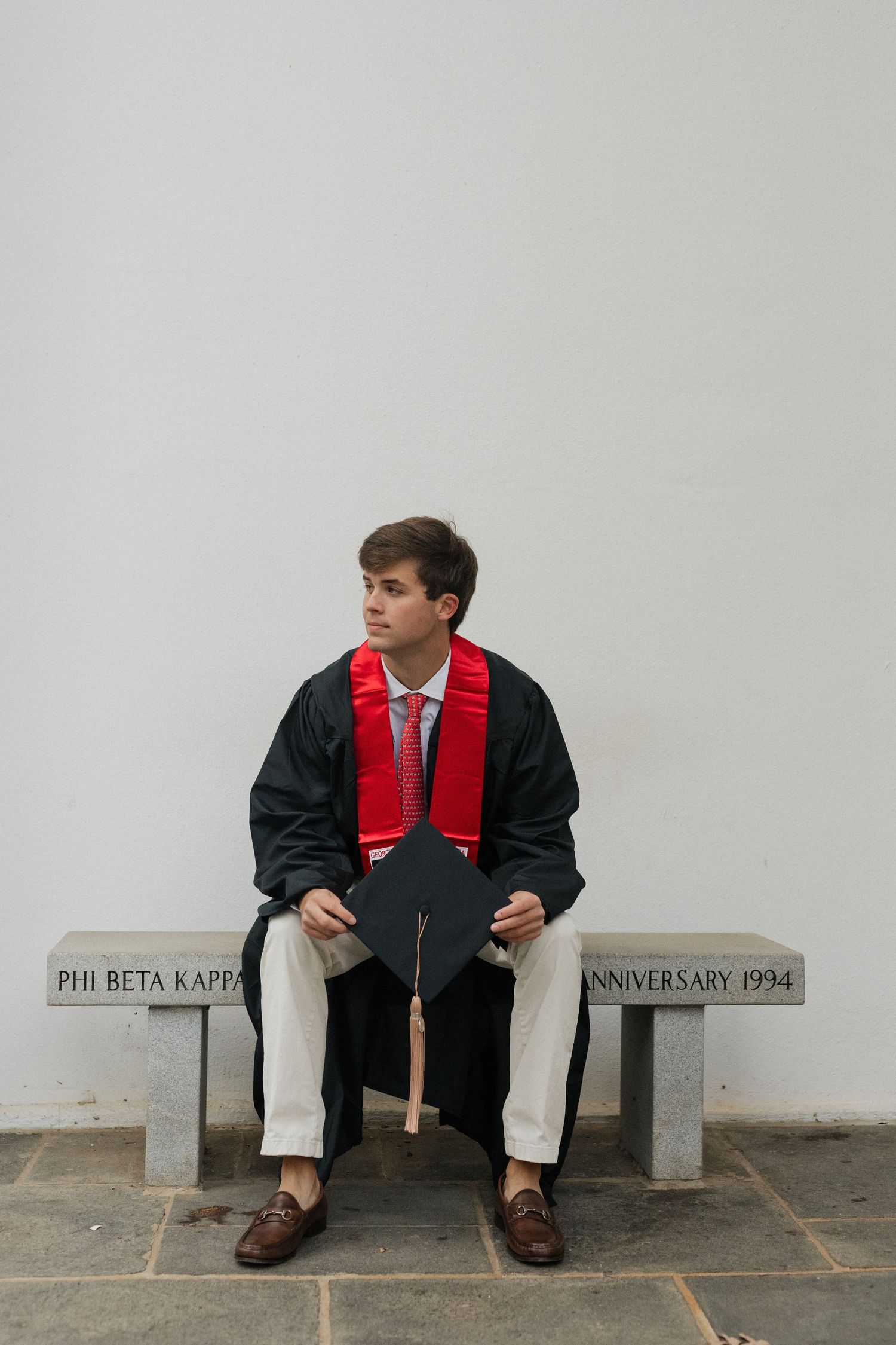 Graduate in red and black regalia sits on stone bench marked with Phi Beta Kappa inscription.