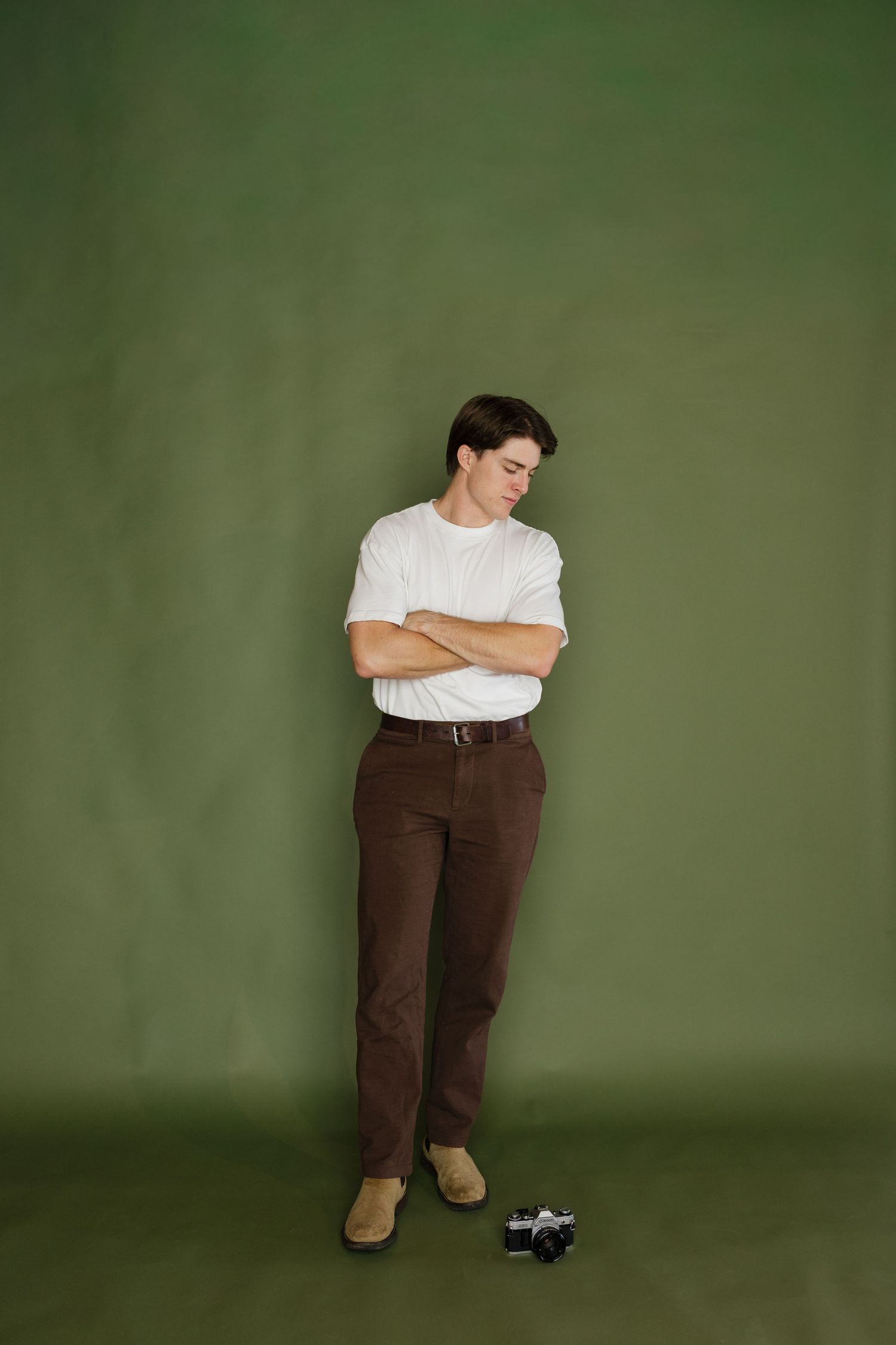 Young man in white t-shirt and brown pants standing with arms crossed against a green studio backdrop.