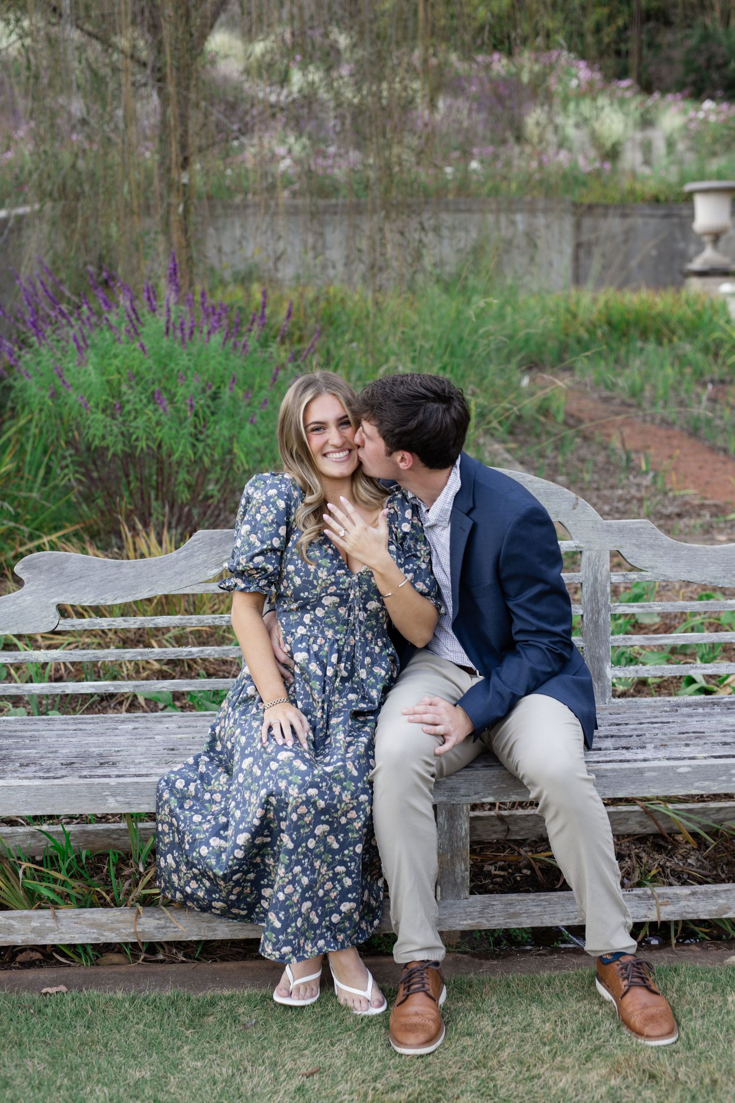 Couple shares intimate moment on wooden bench in outdoor garden setting.