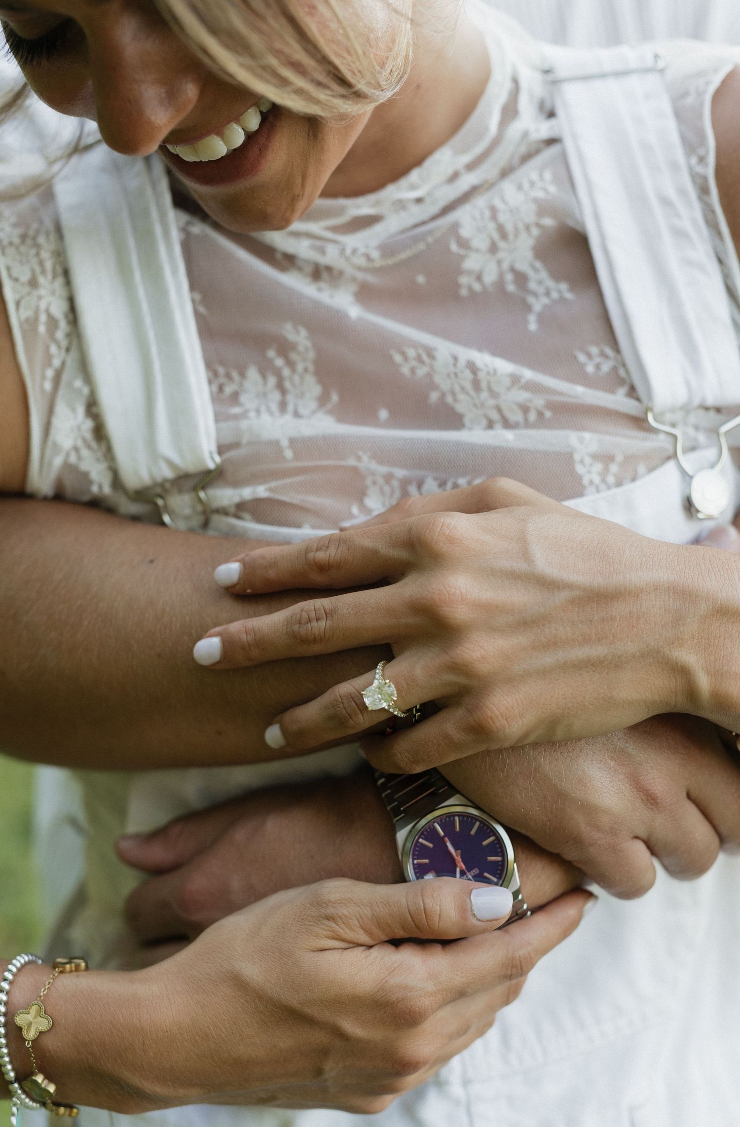 Bride in lace wedding dress smiling while wearing diamond engagement ring, holding hands with partner wearing a classic watch.
