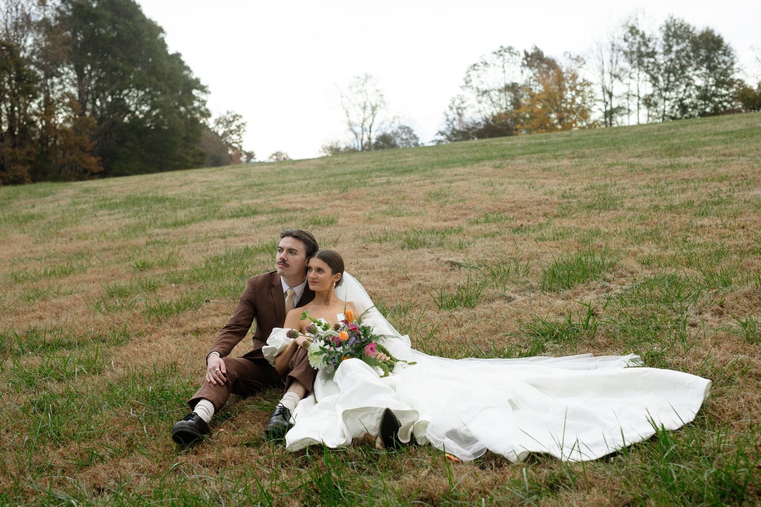 Wedding couple poses dramatically on grassy hillside with flowing white dress.