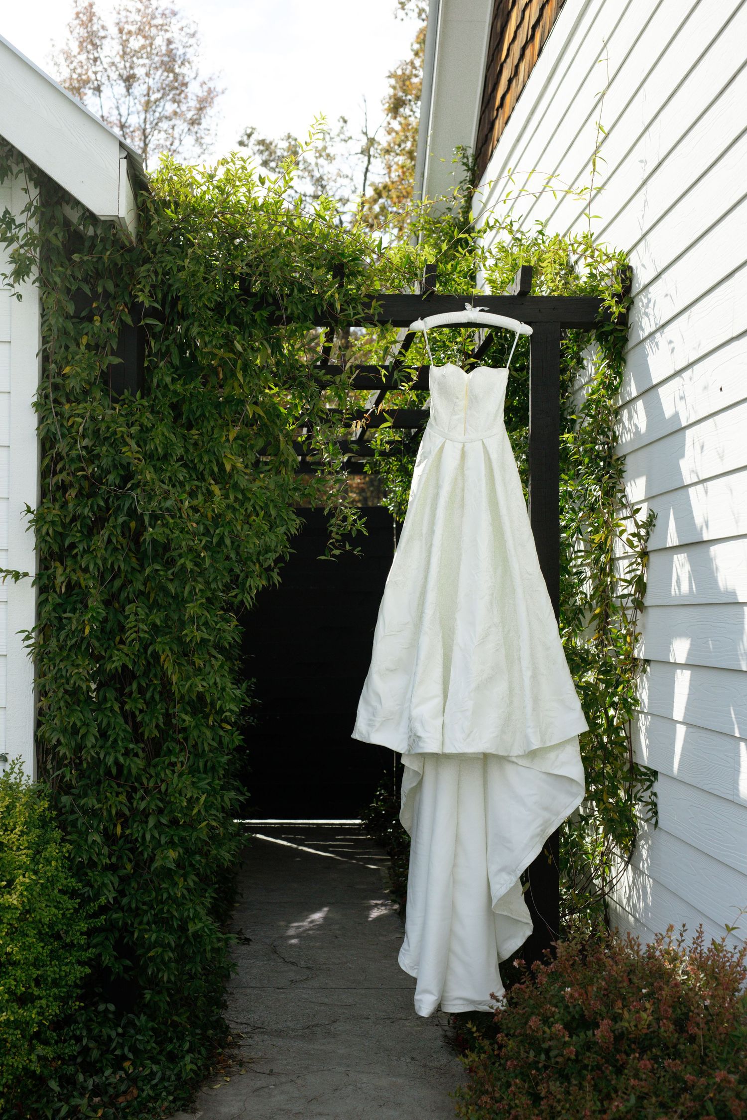 White wedding dress displayed hanging outdoors against white siding and green foliage.