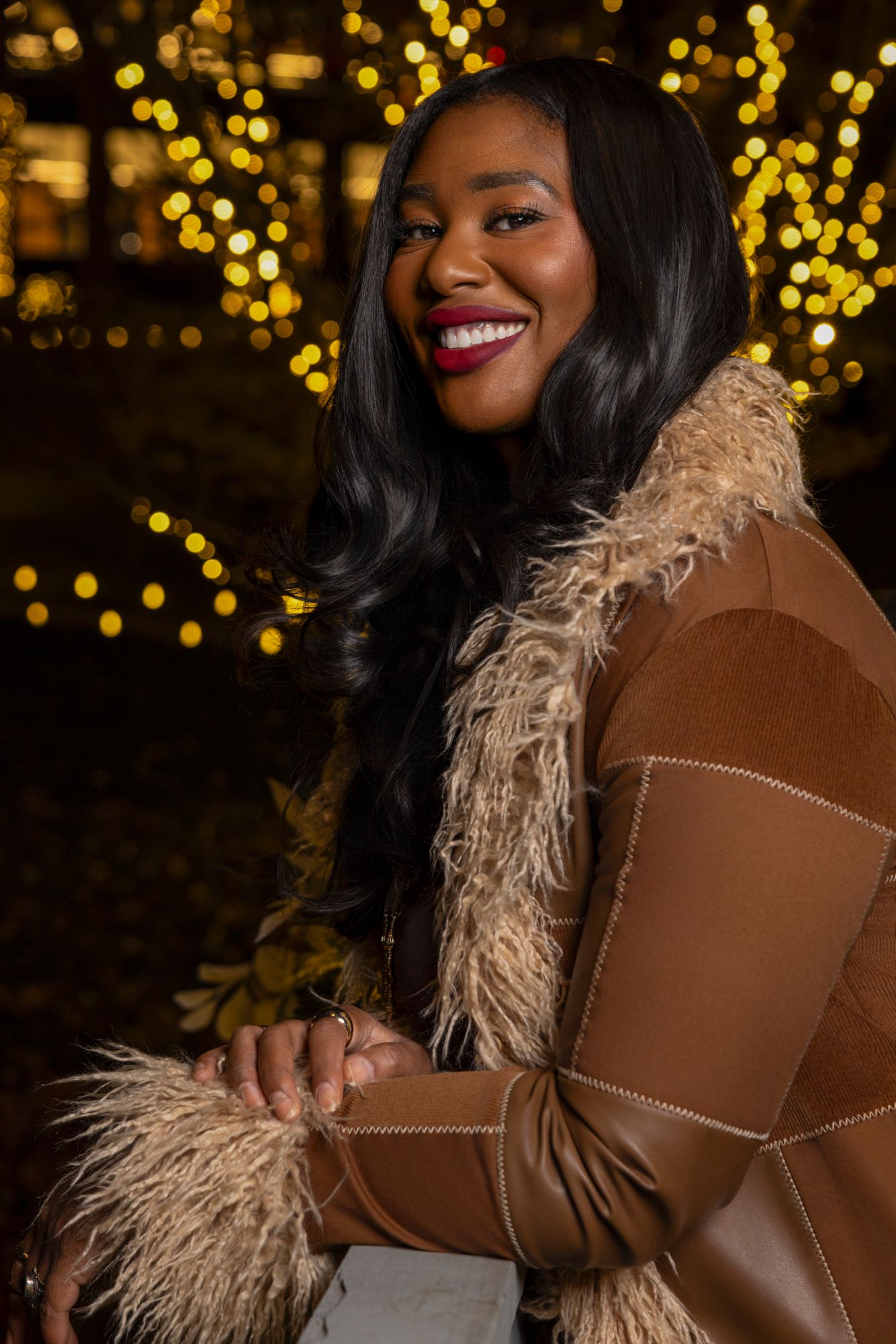 Stylish winter outfit featuring a brown coat with fur trim against festive holiday lights.