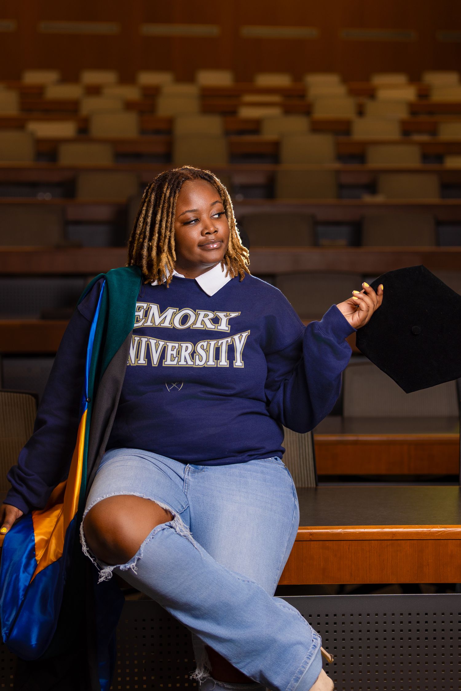 Student celebrates in Emory University sweatshirt and graduation cap while sitting in lecture hall.