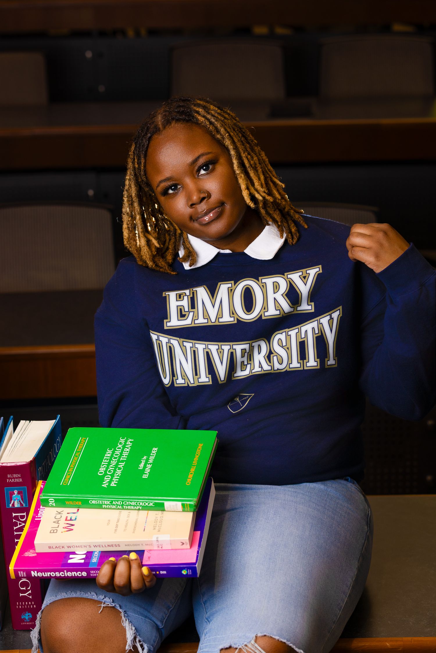 Student holds textbooks while wearing Emory University sweatshirt in classroom amphitheater.