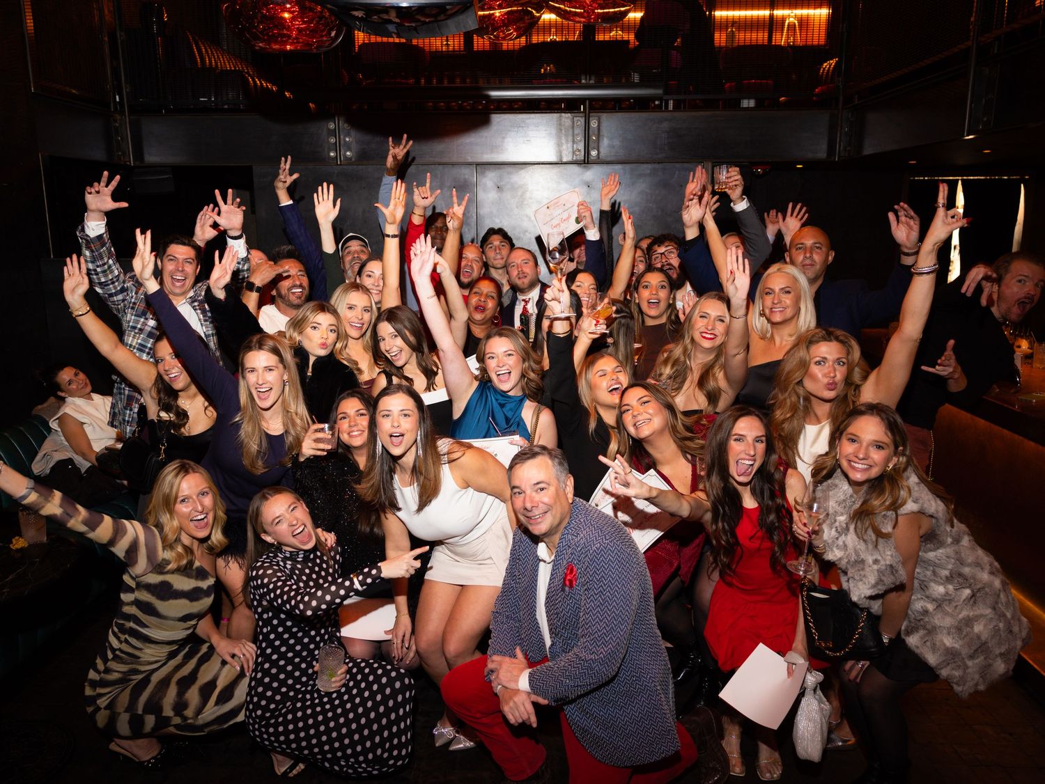 Large crowd of party-goers celebrating with raised arms in dimly lit venue.