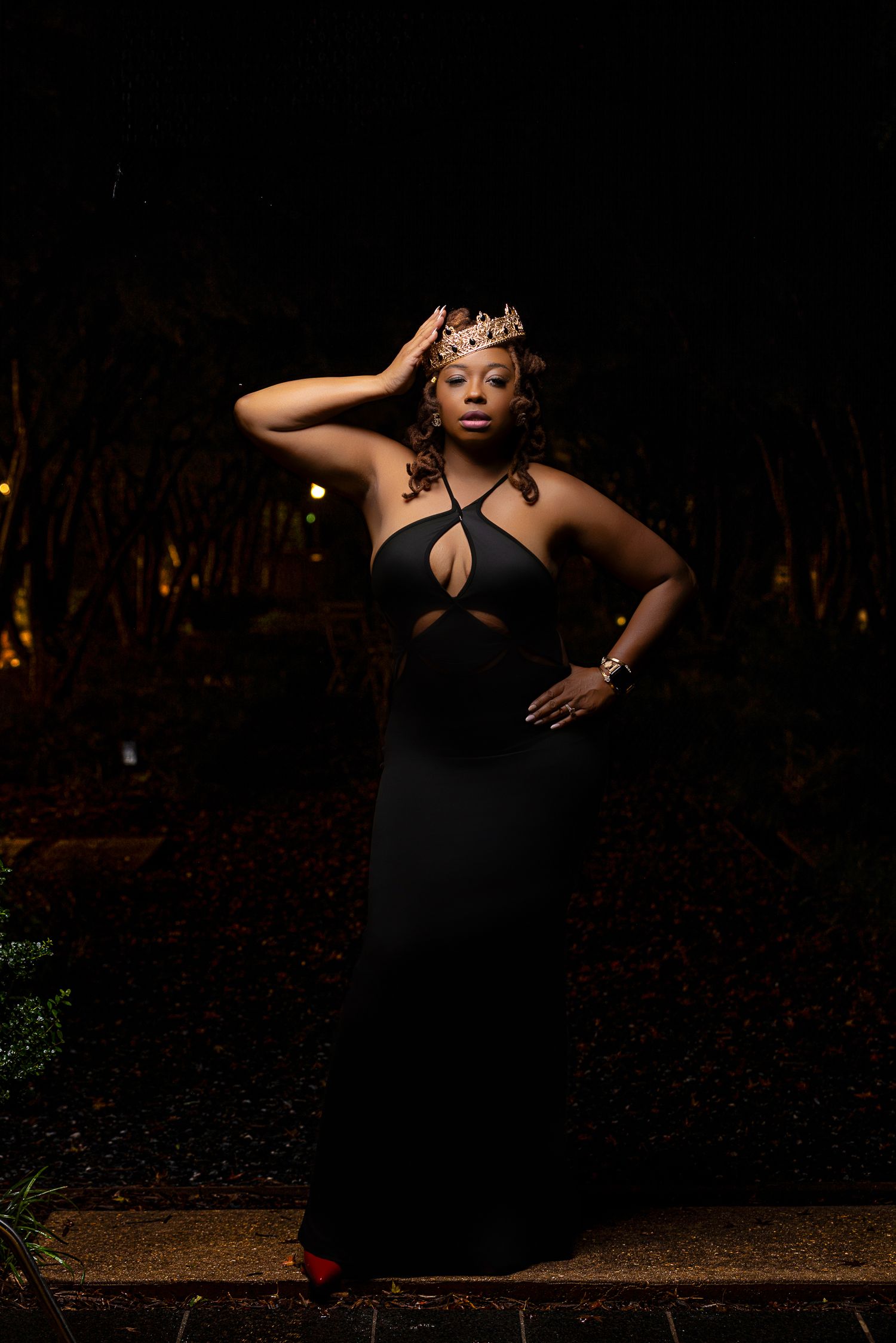 Dramatic evening portrait in black cutout dress and crown against dark outdoor setting.