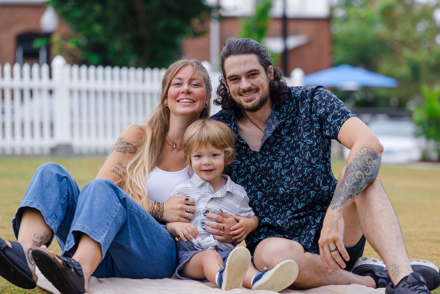 Family lying together on blanket outdoors with white picket fence in background.