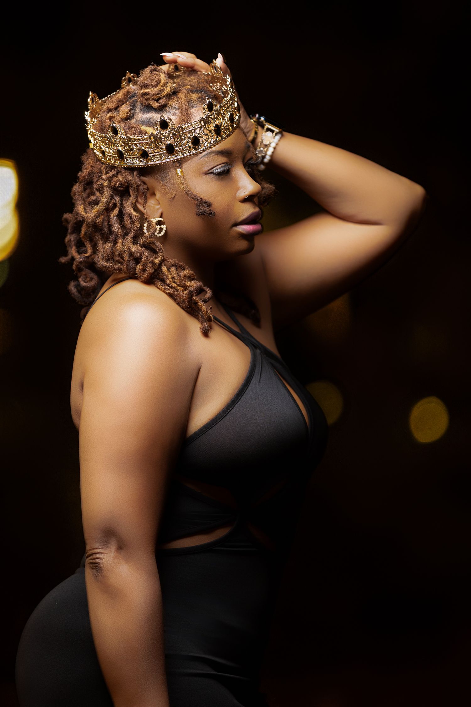 Dramatic portrait wearing ornate gold crown against dark background with bokeh lights.