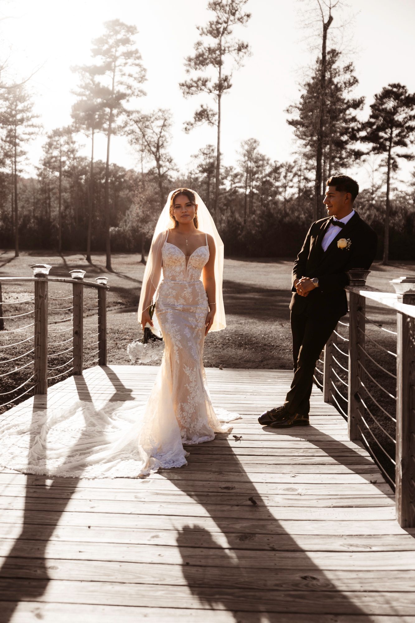 A couple in wedding attire stands on a wooden bridge during golden hour.