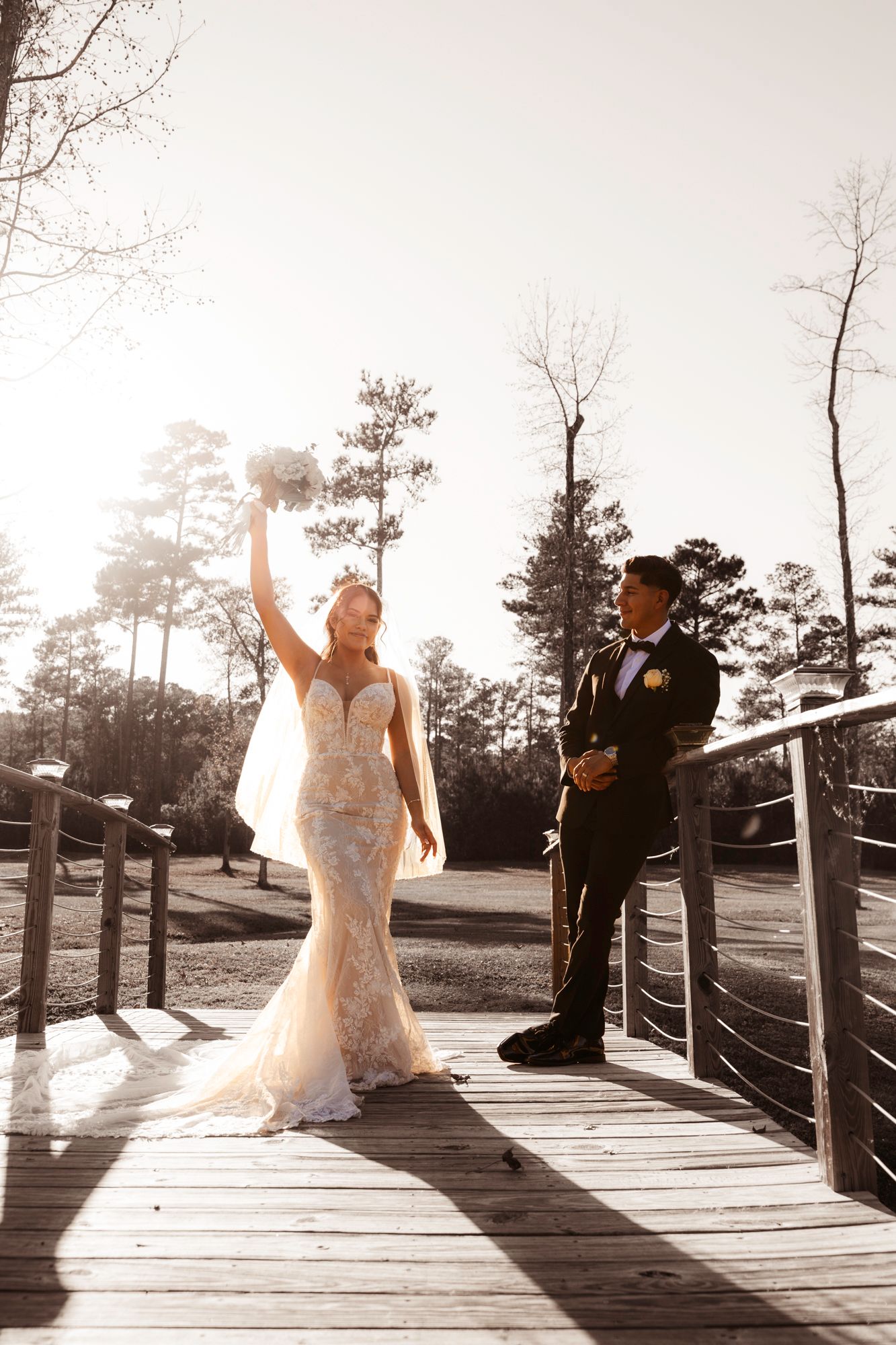 Bride raises bouquet joyfully while standing with groom on wooden deck during sunset.