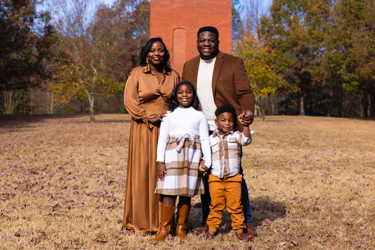 Family of four poses together for portrait in front of brick building during autumn.