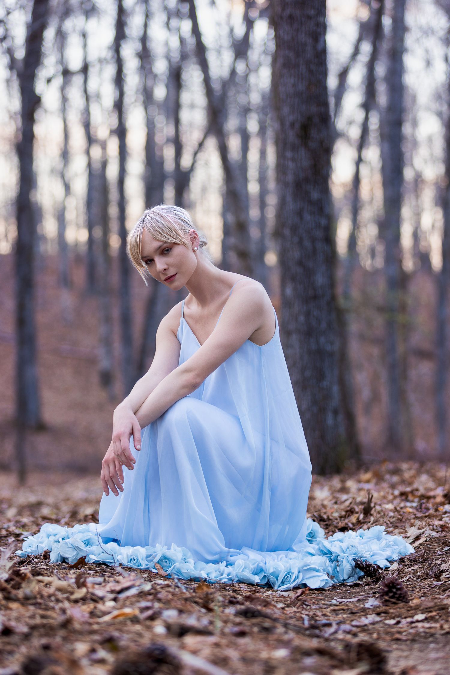 Dreamy portrait in pale blue flowing gown among fallen autumn leaves in forest.