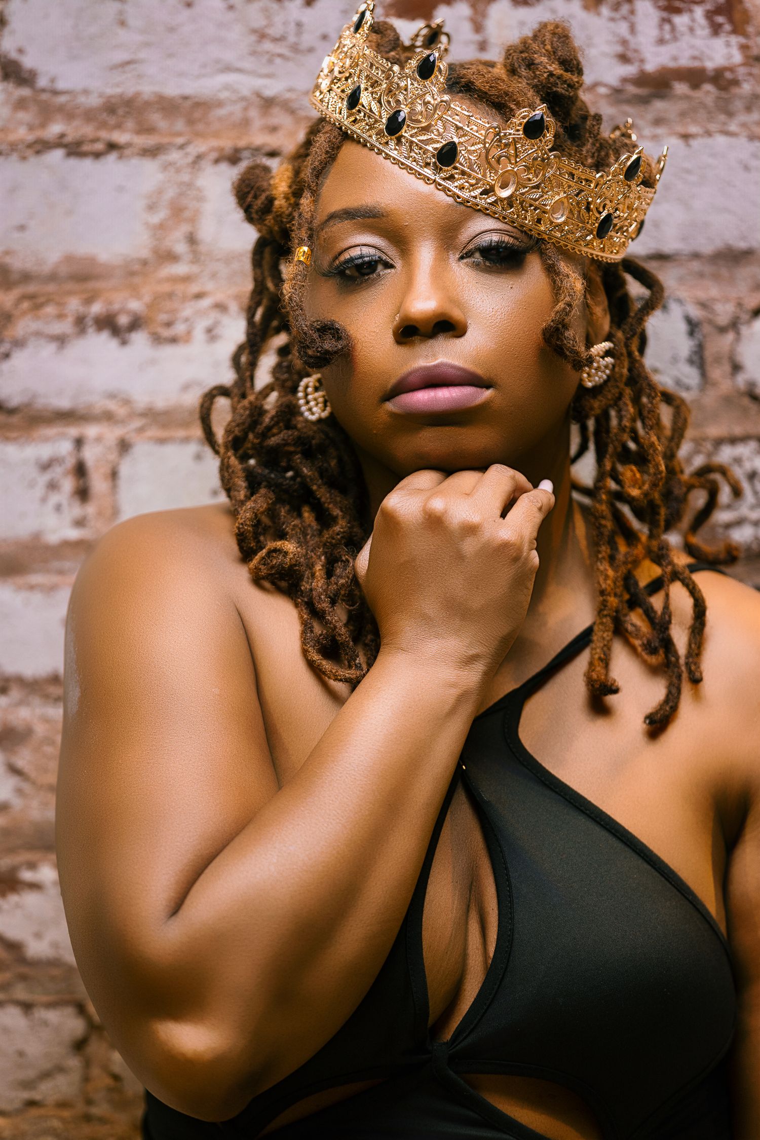 Artistic portrait with gold crown and natural locs against textured brick wall.