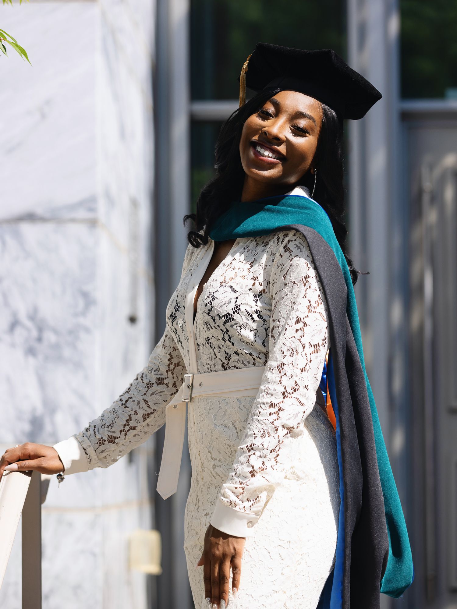 Graduate in white lace gown and teal stole stands against marble wall for graduation photos.