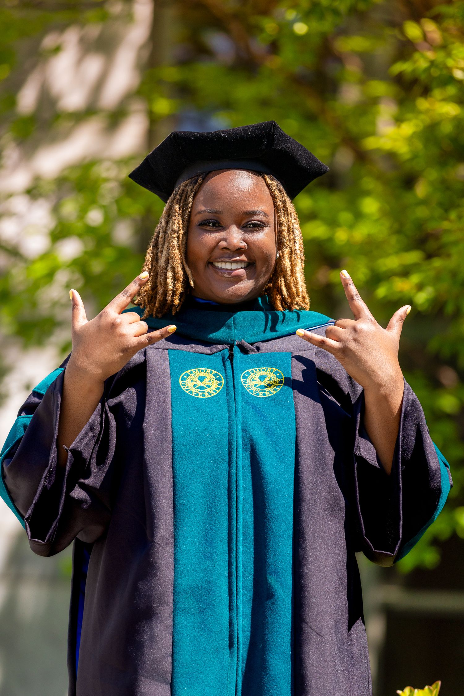 Graduate in teal and purple academic regalia gives thumbs up gesture while celebrating outdoors.