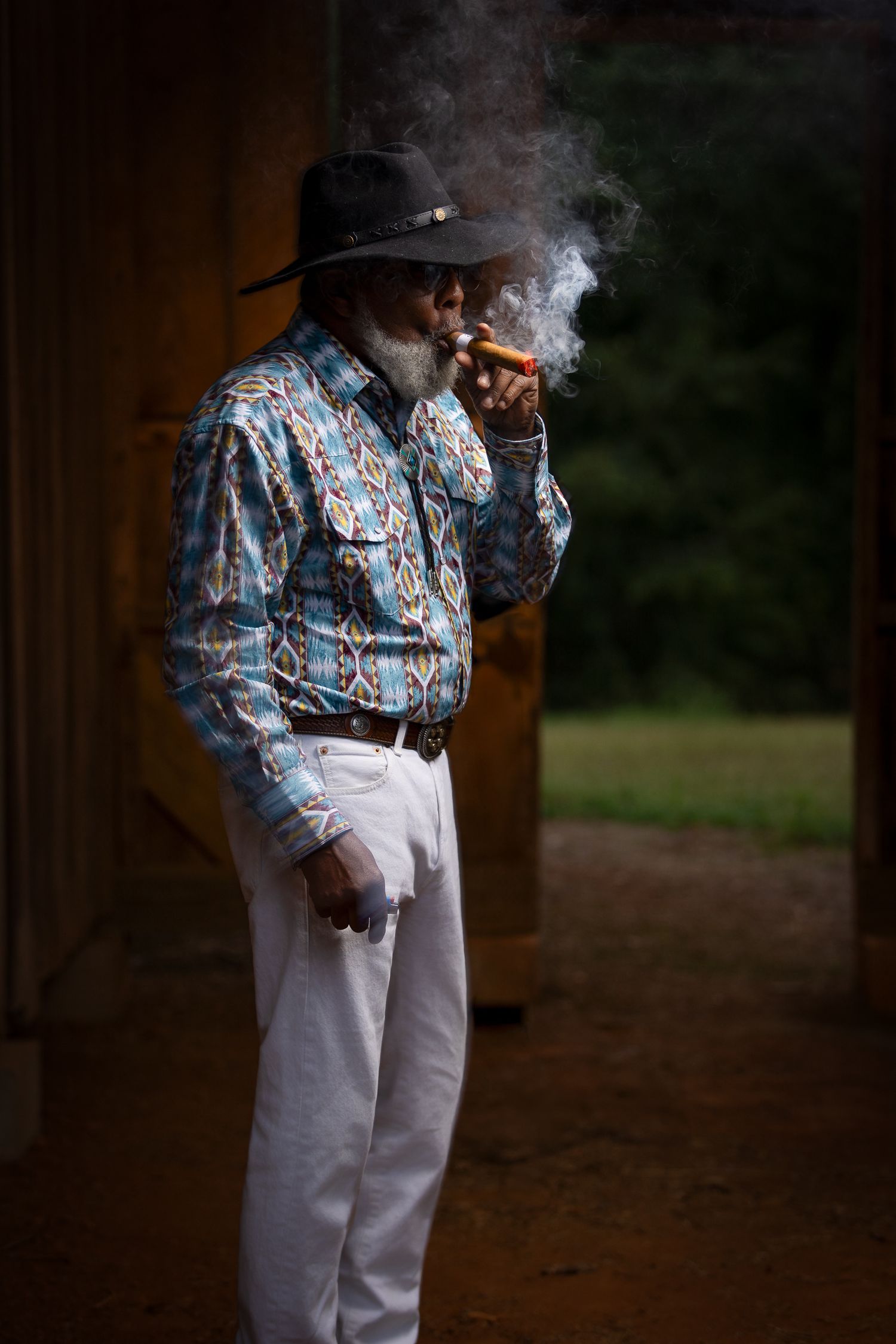 Person in western attire smoking a cigar with dramatic lighting in barn setting.