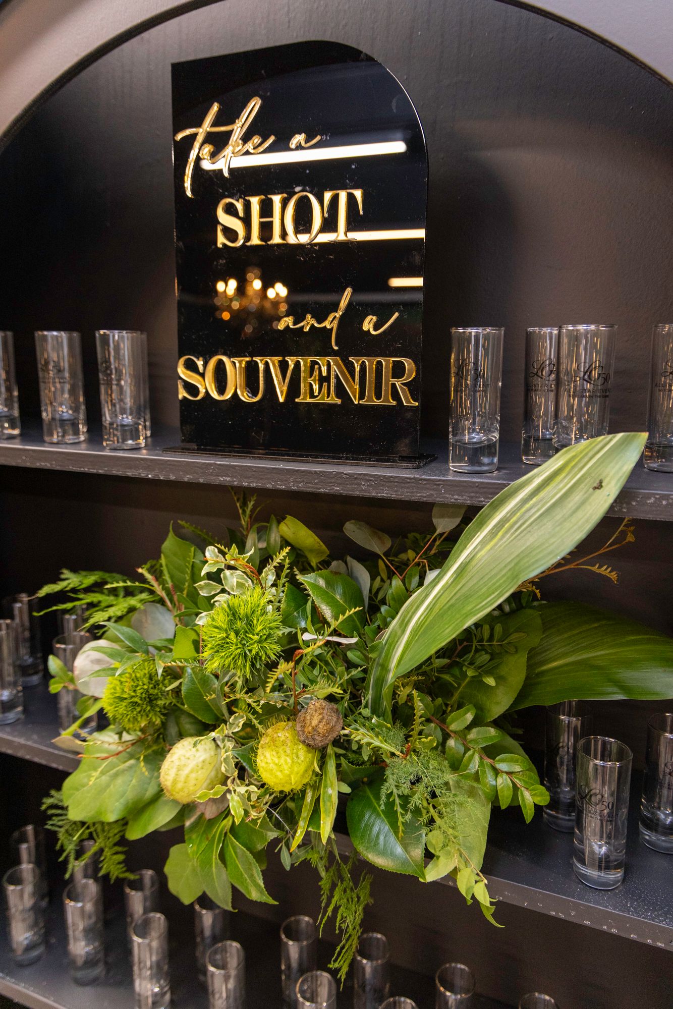 Black wall-mounted shelving unit with shot glasses and fresh green floral arrangements.