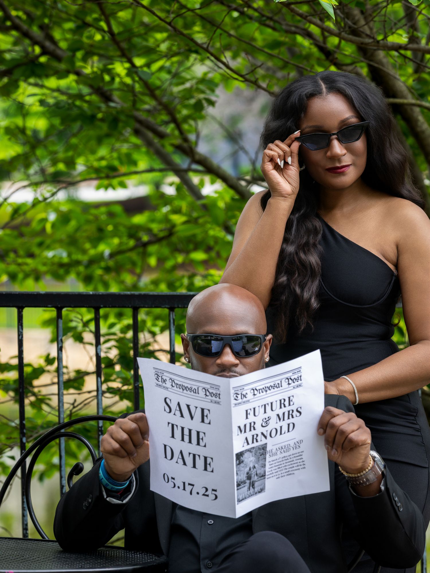 A couple creates a creative save-the-date photo with sunglasses and a newspaper prop.