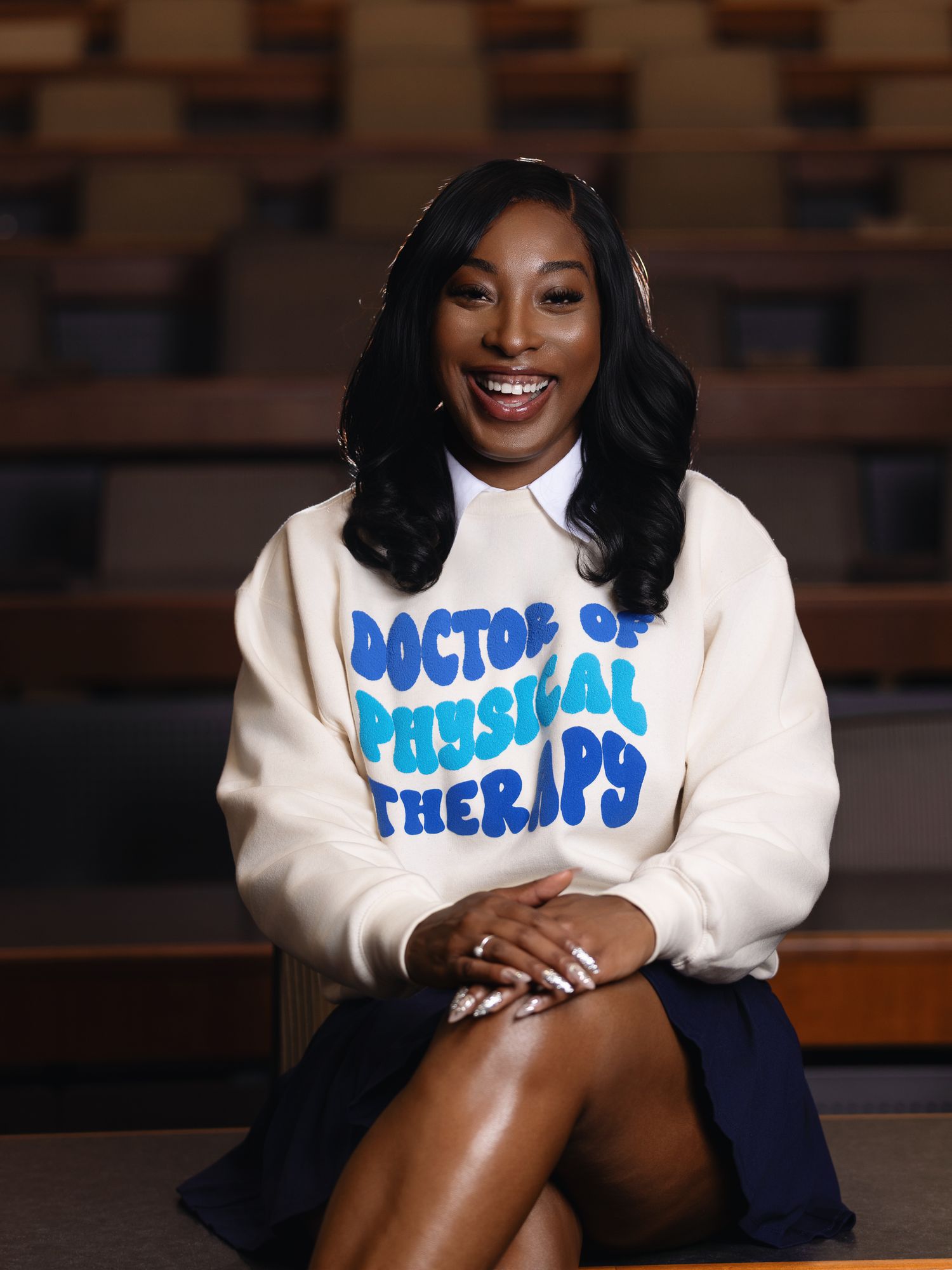 Graduate wearing white sweatshirt with blue text sitting in auditorium setting.