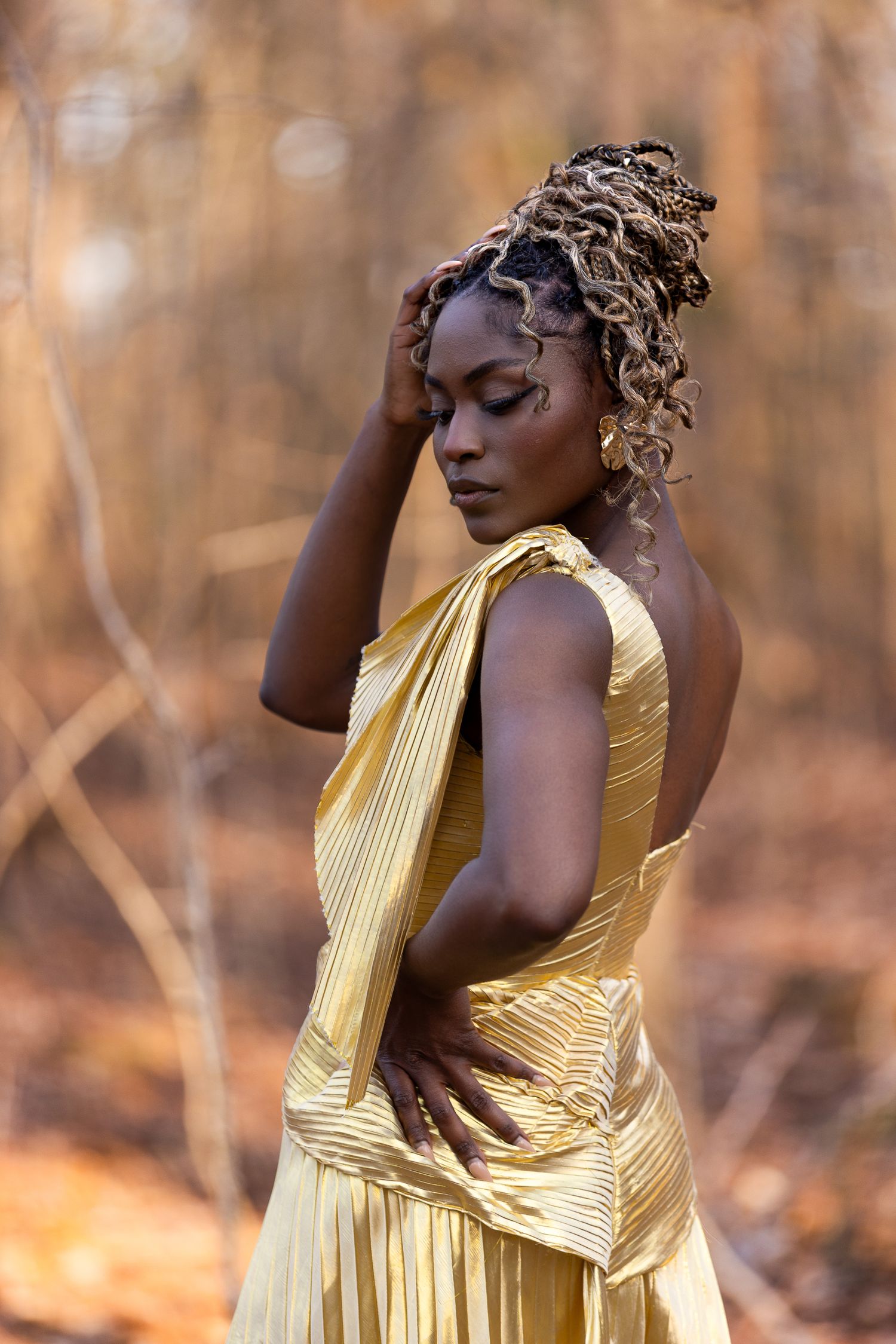 Model in flowing gold dress poses elegantly against autumn forest backdrop.