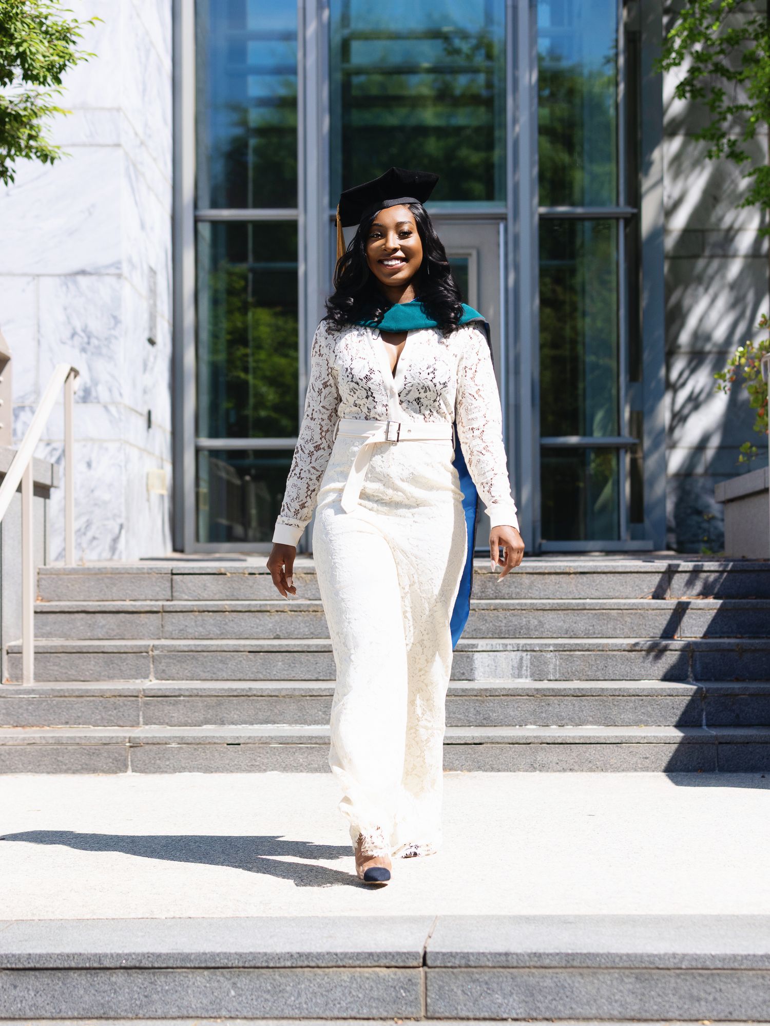 Graduate in elegant white lace dress poses on stone steps in front of glass building entrance.