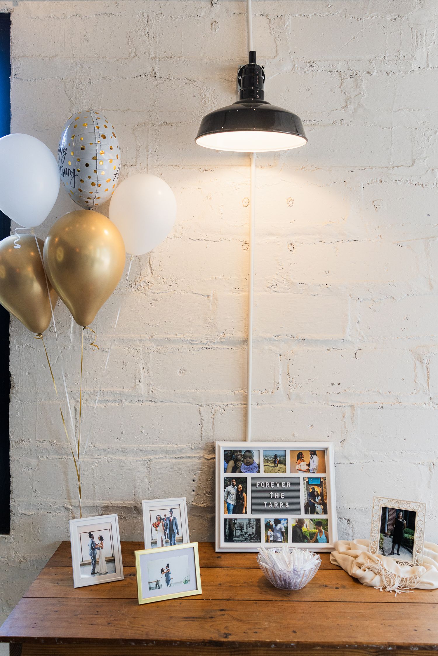 Decorative table setup with photo frames, balloons, and pendant lighting against white brick wall.