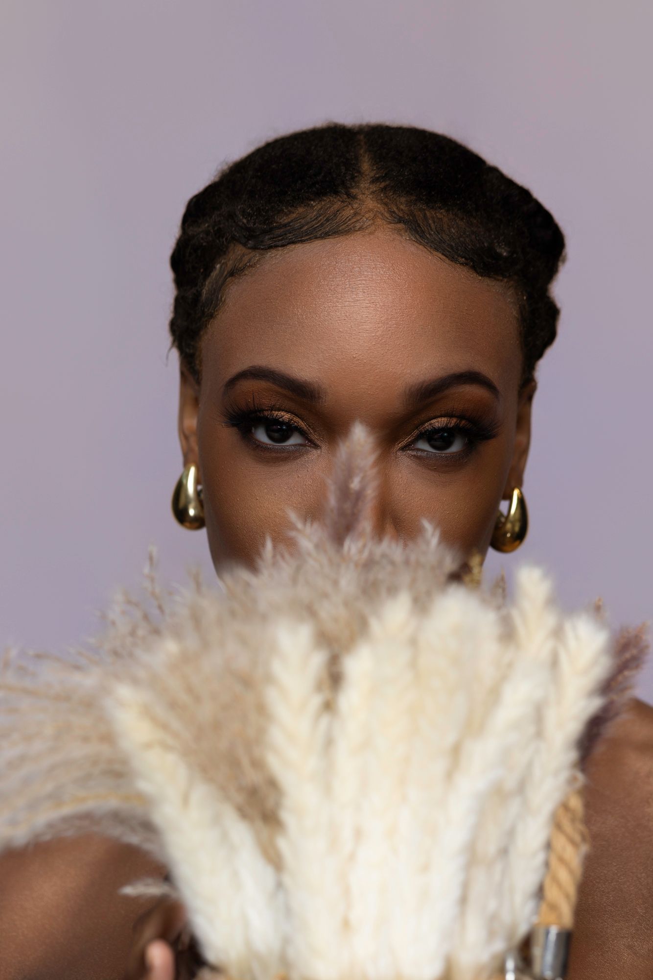 Dramatic beauty shot with cream feather fan and gold earrings against purple backdrop.