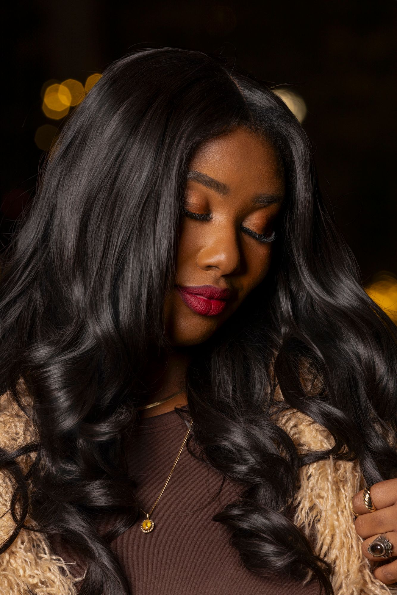 Fashion portrait of a woman in brown sweater with wavy black hair against golden bokeh lights.