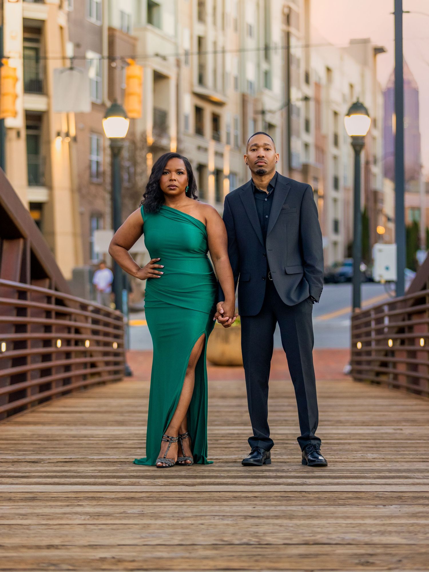 A couple poses on a wooden bridge, with one wearing an elegant green dress and the other in a dark suit.