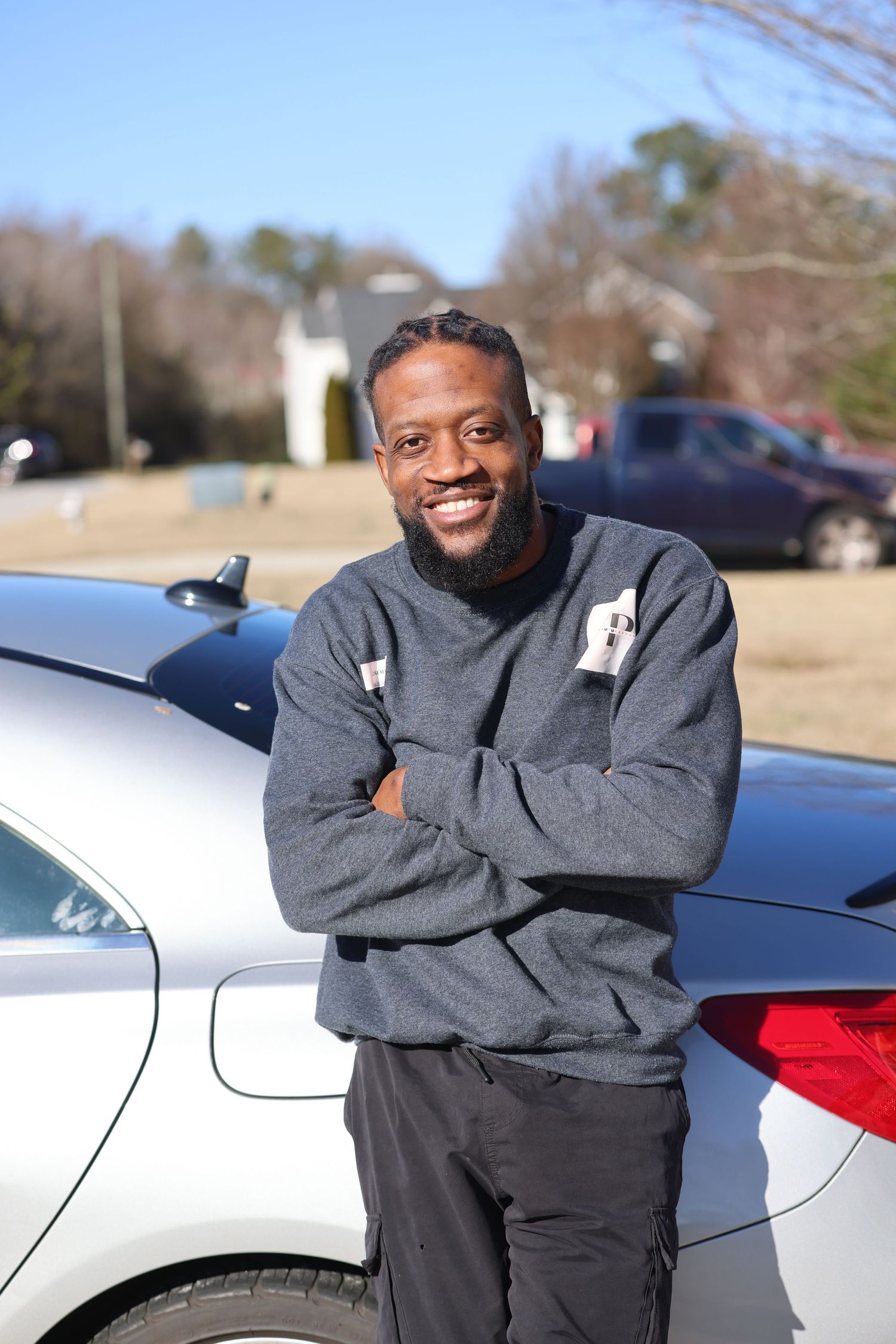 Person in grey sweater smiling and posing casually by parked cars.