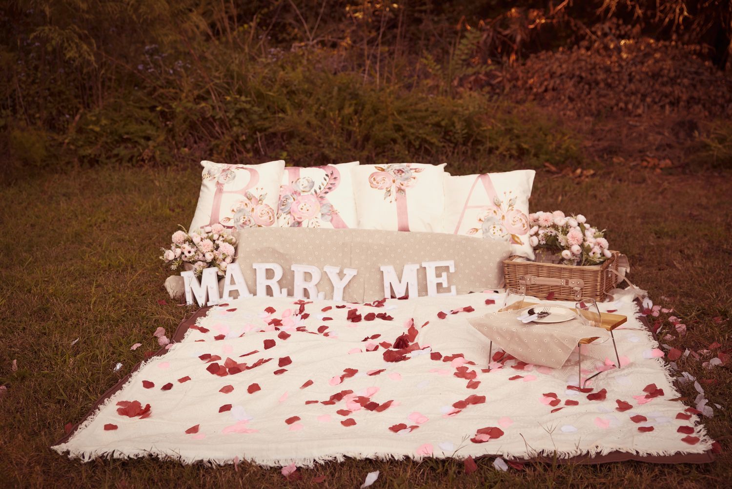 Romantic proposal setup with 'Marry Me' sign and rose petals on white fabric.