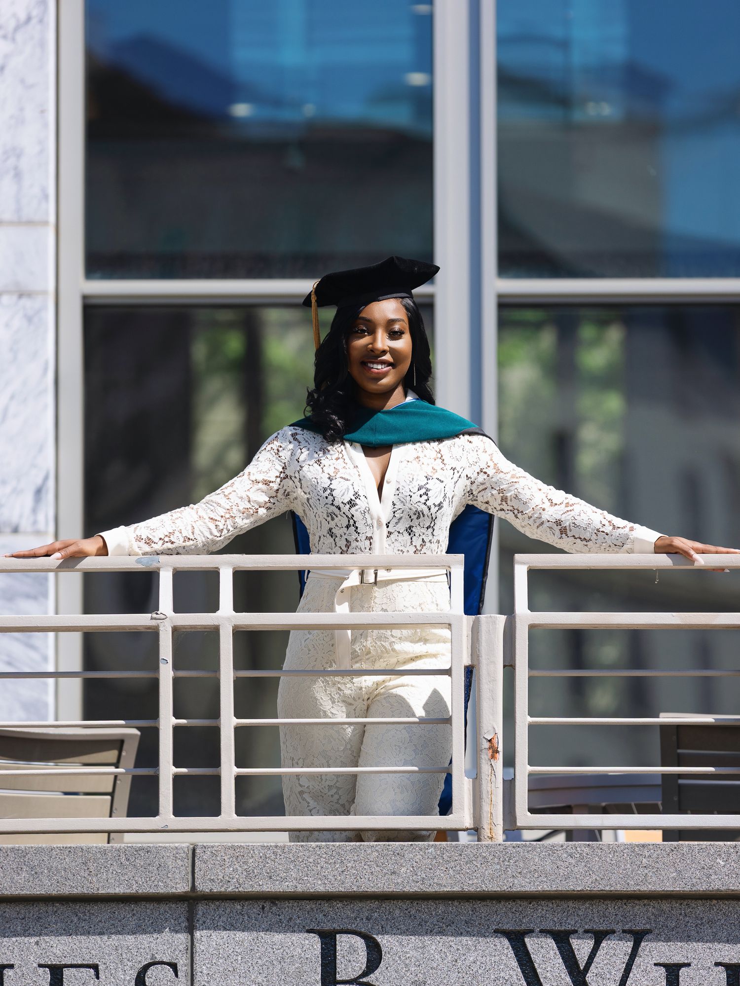 Graduate posing on a modern building balcony in academic regalia.