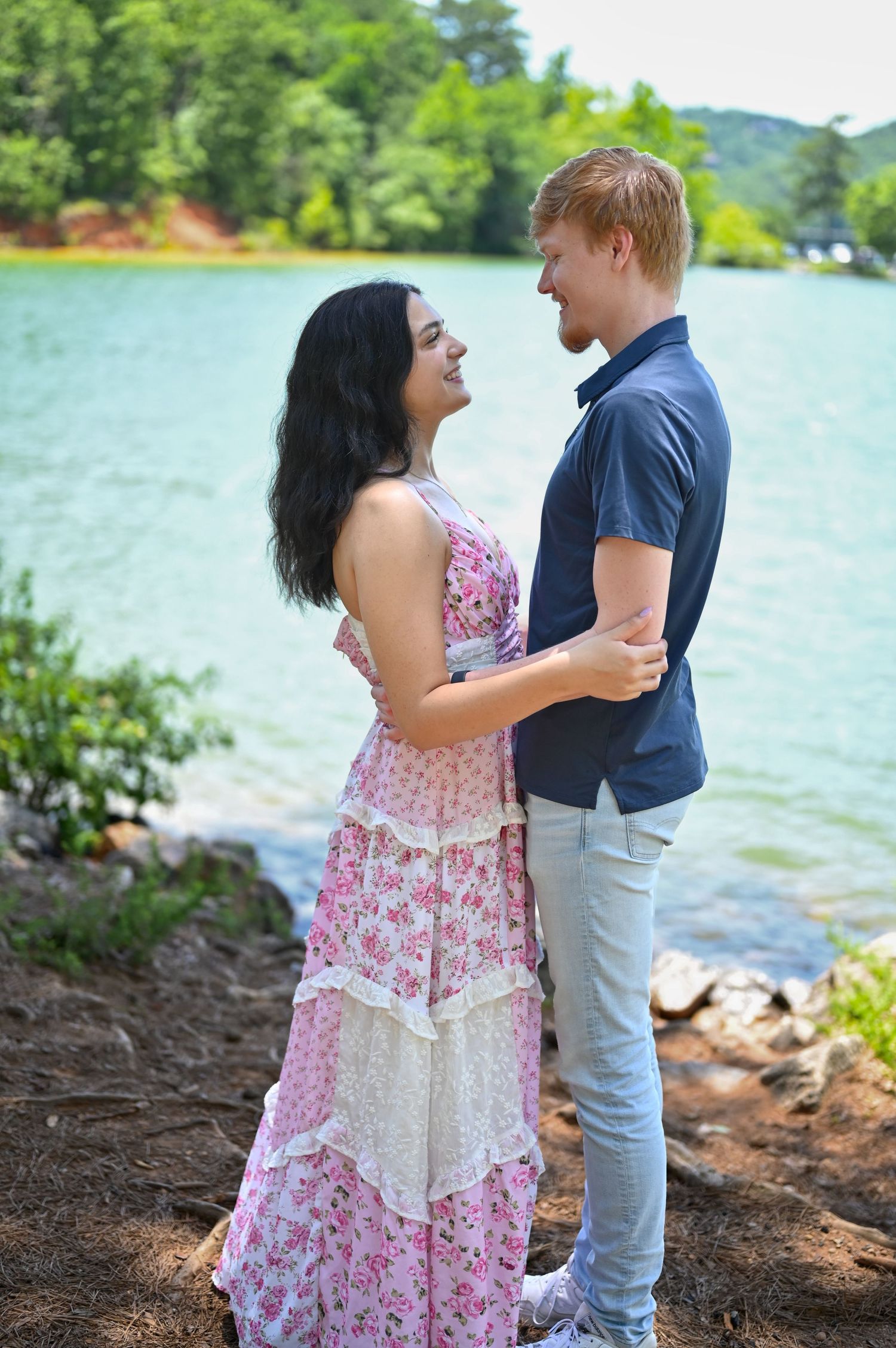 Couple smiling at each other in a romantic embrace by a serene lake surrounded by lush green trees.