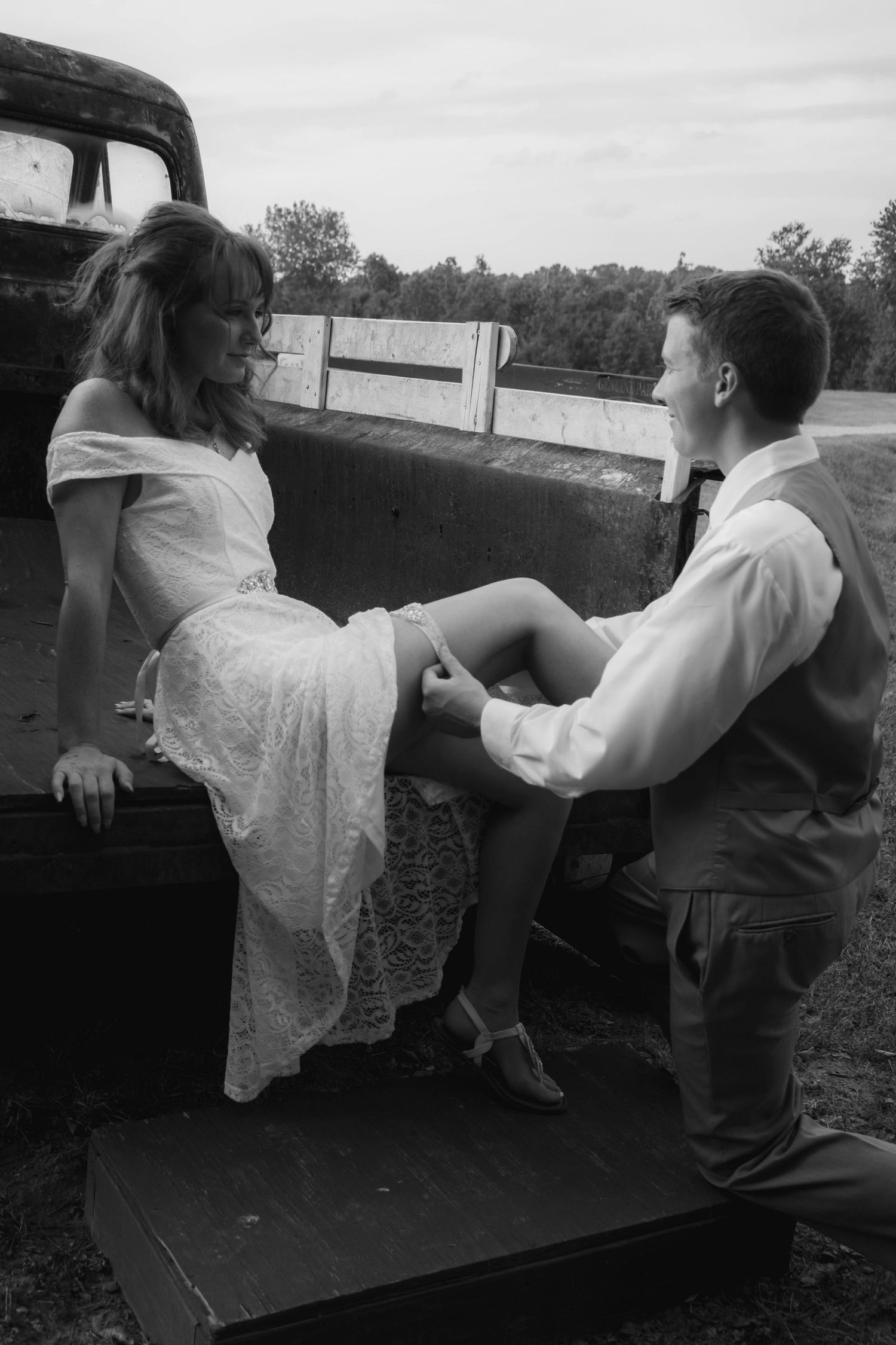 Black and white photo of groom kneeling before bride seated on vintage truck tailgate.