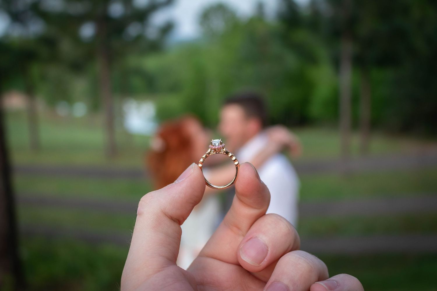 Close-up of a diamond engagement ring held up with a couple embracing in the background.