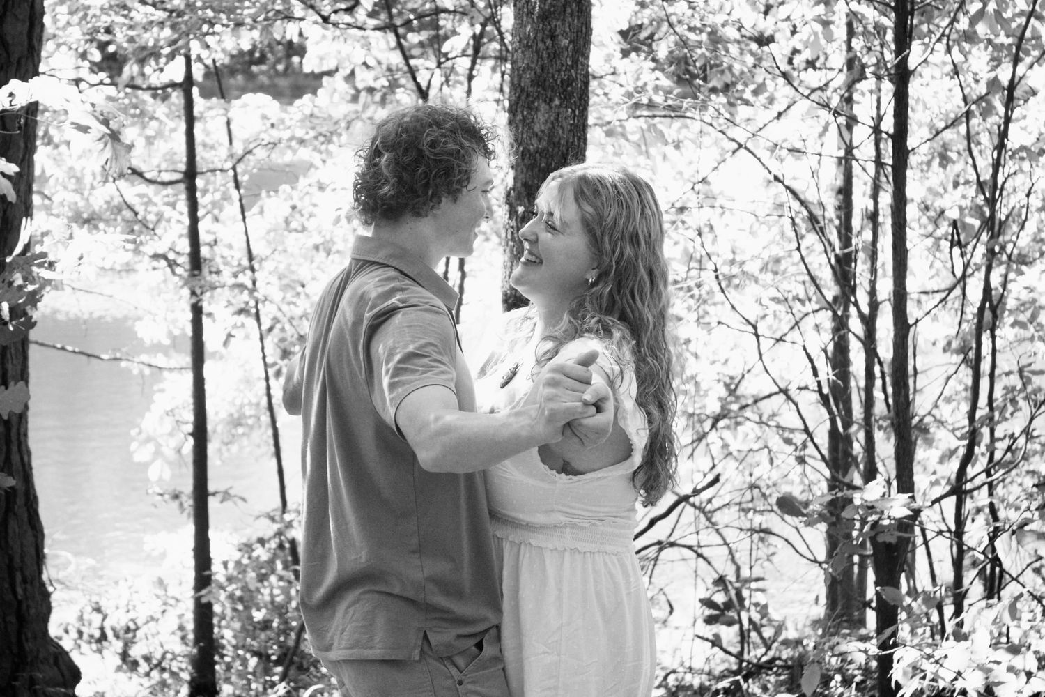 Black and white photo of a romantic couple dancing and smiling together in a forest setting.