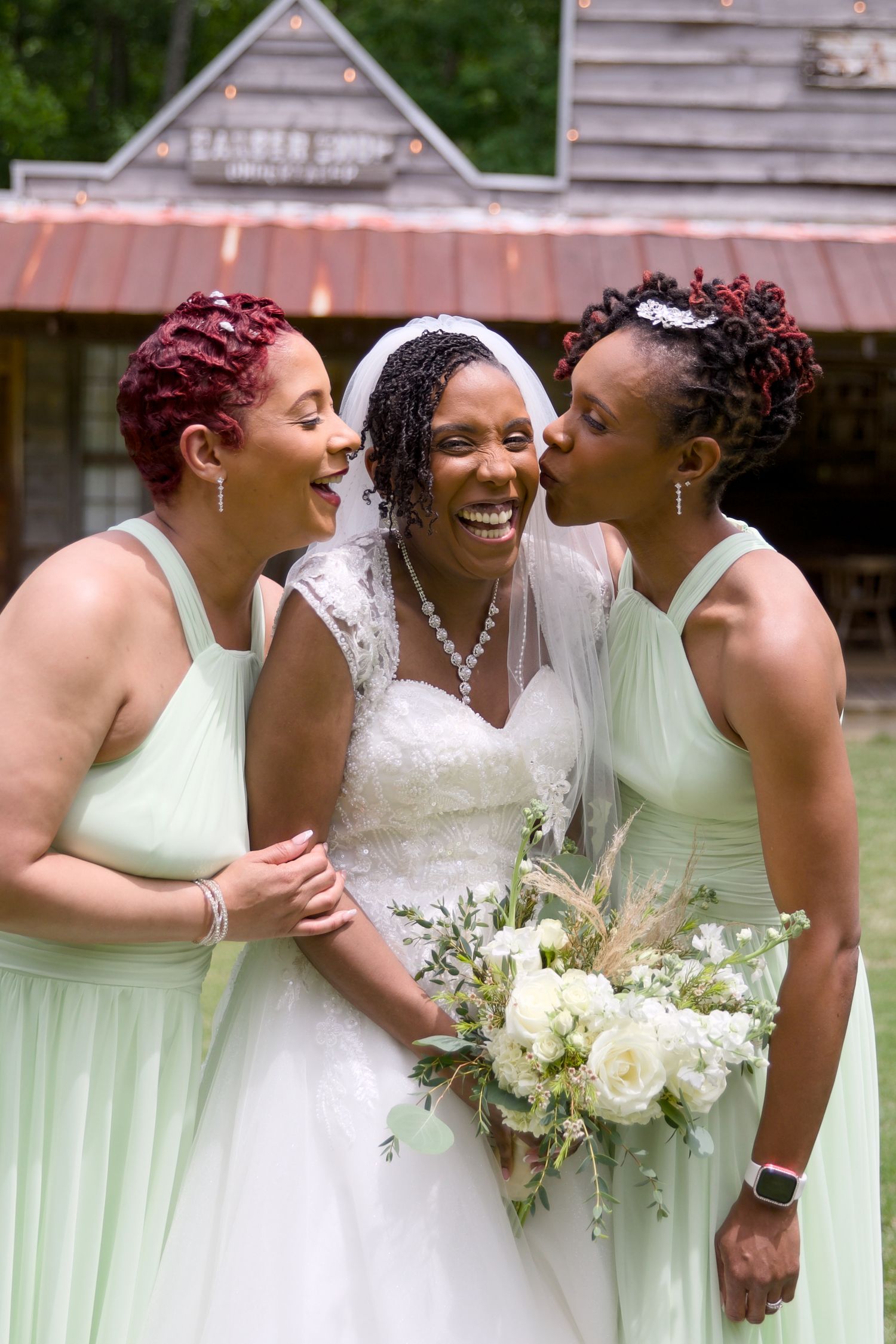 Bride in white gown and veil smiles as two bridesmaids in mint green dresses kiss her cheeks outdoors.