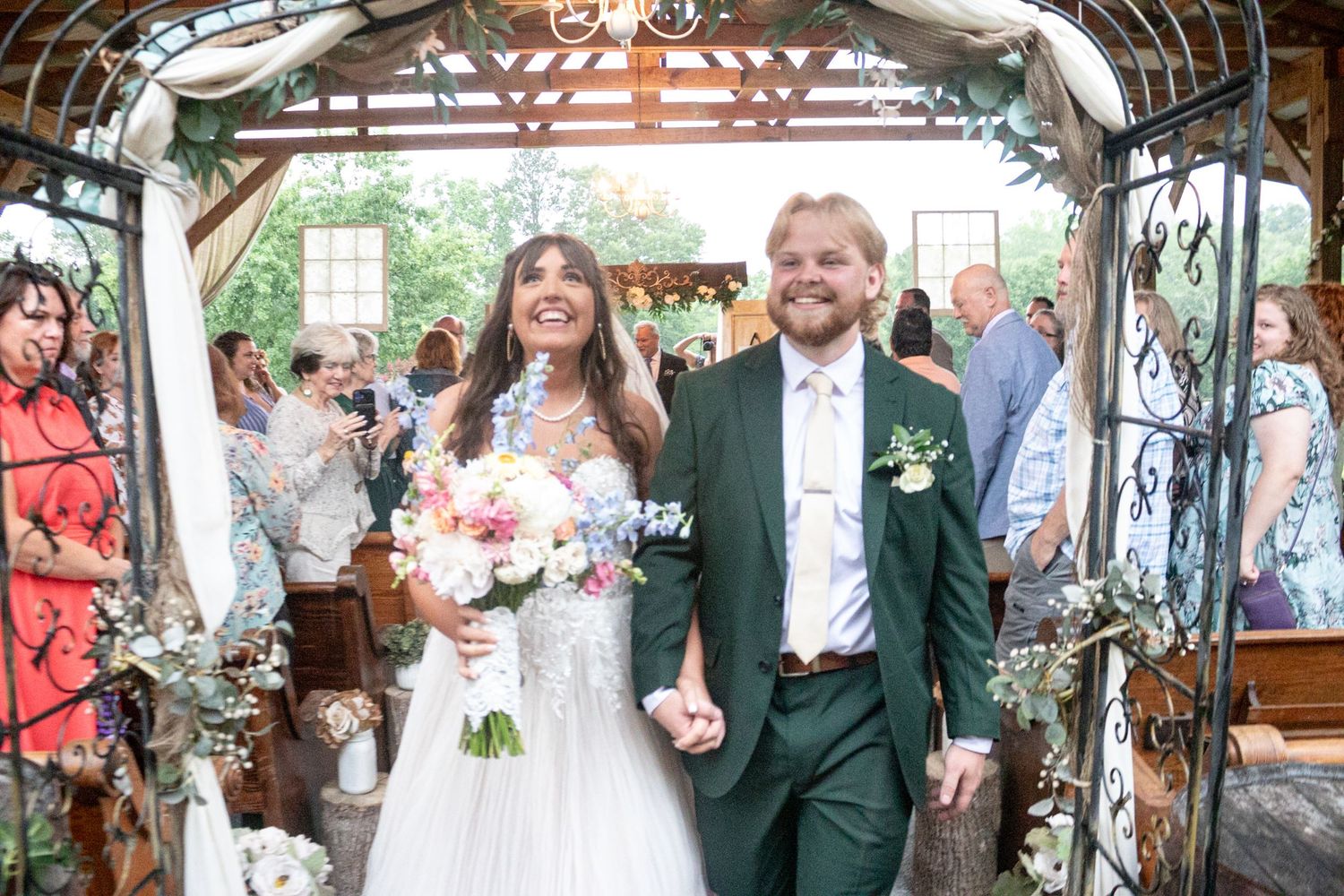 Smiling newlyweds walking hand in hand through ceremony exit, bride holding colorful bouquet, groom in green suit.