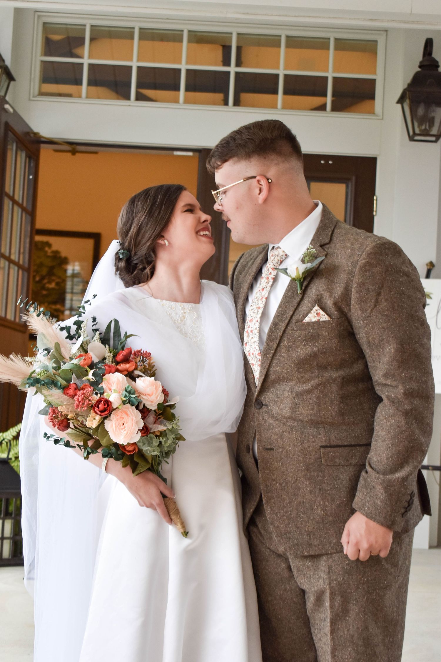 Bride and groom share a romantic moment outdoors, she holds autumn bouquet in white gown, he wears brown tweed suit.