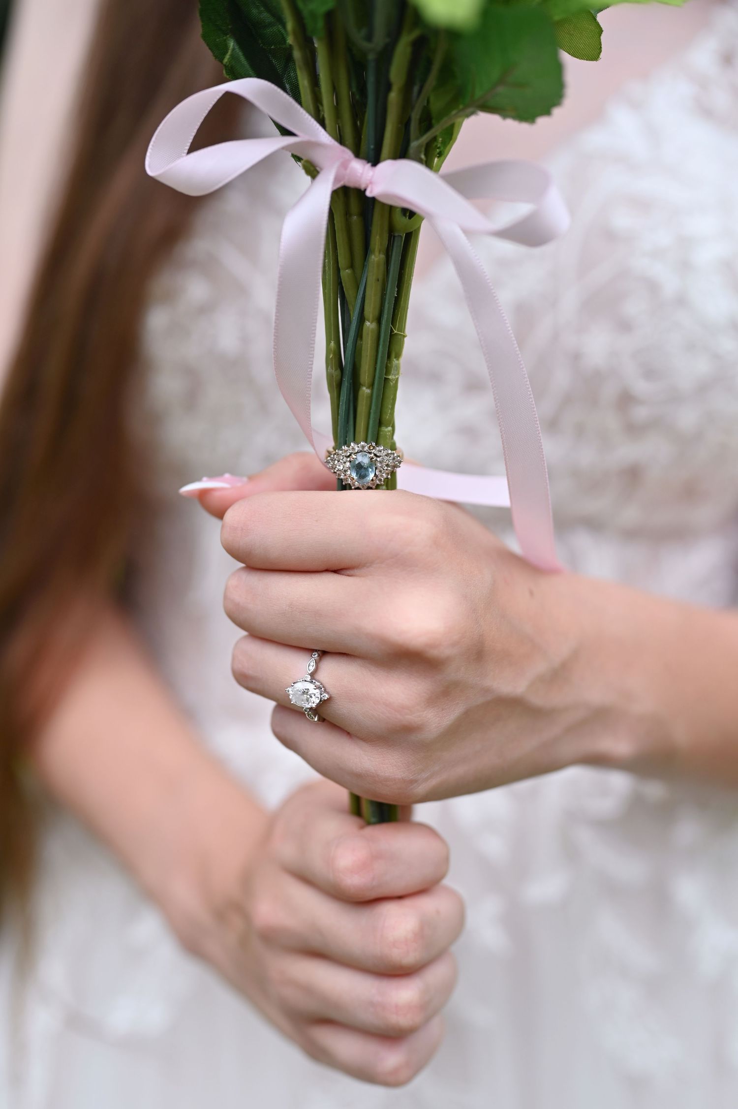 Bride holding a floral bouquet tied with pink ribbon, showcasing a diamond engagement ring on her finger.