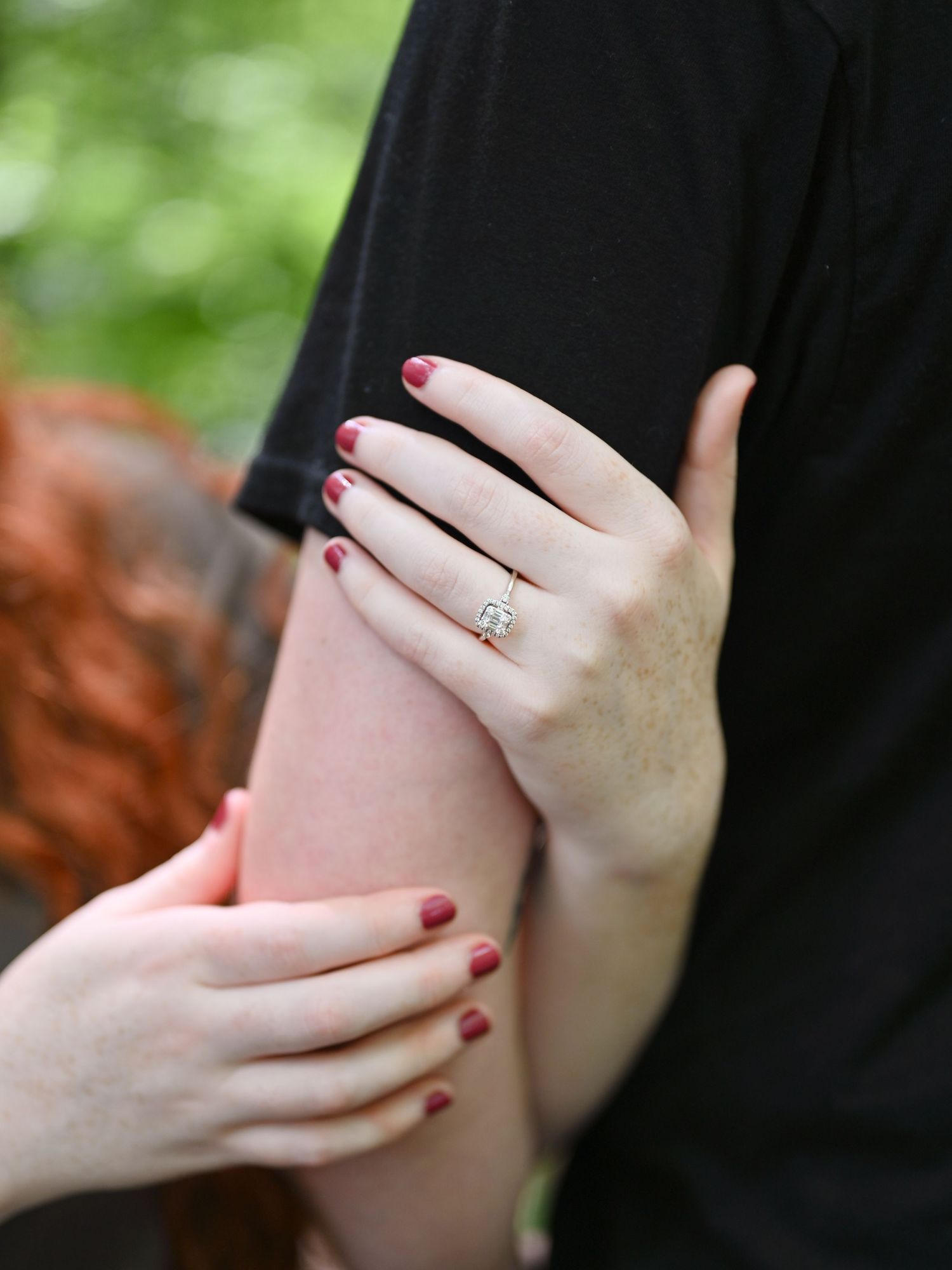 Close-up of a woman's hand with red nails and a diamond engagement ring resting on a man's arm.