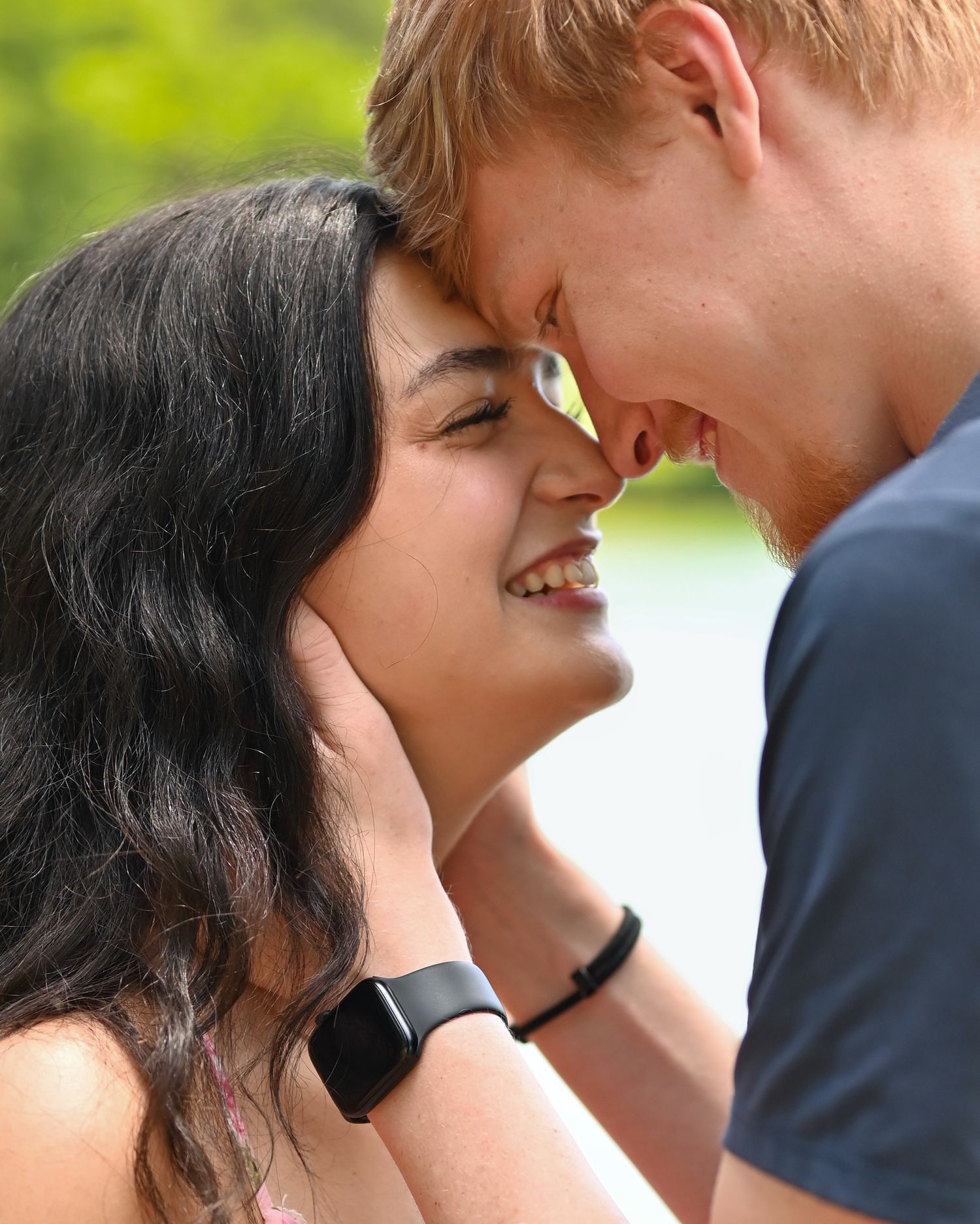 A smiling young couple touching foreheads romantically outdoors, the woman wearing a smartwatch and floral dress.