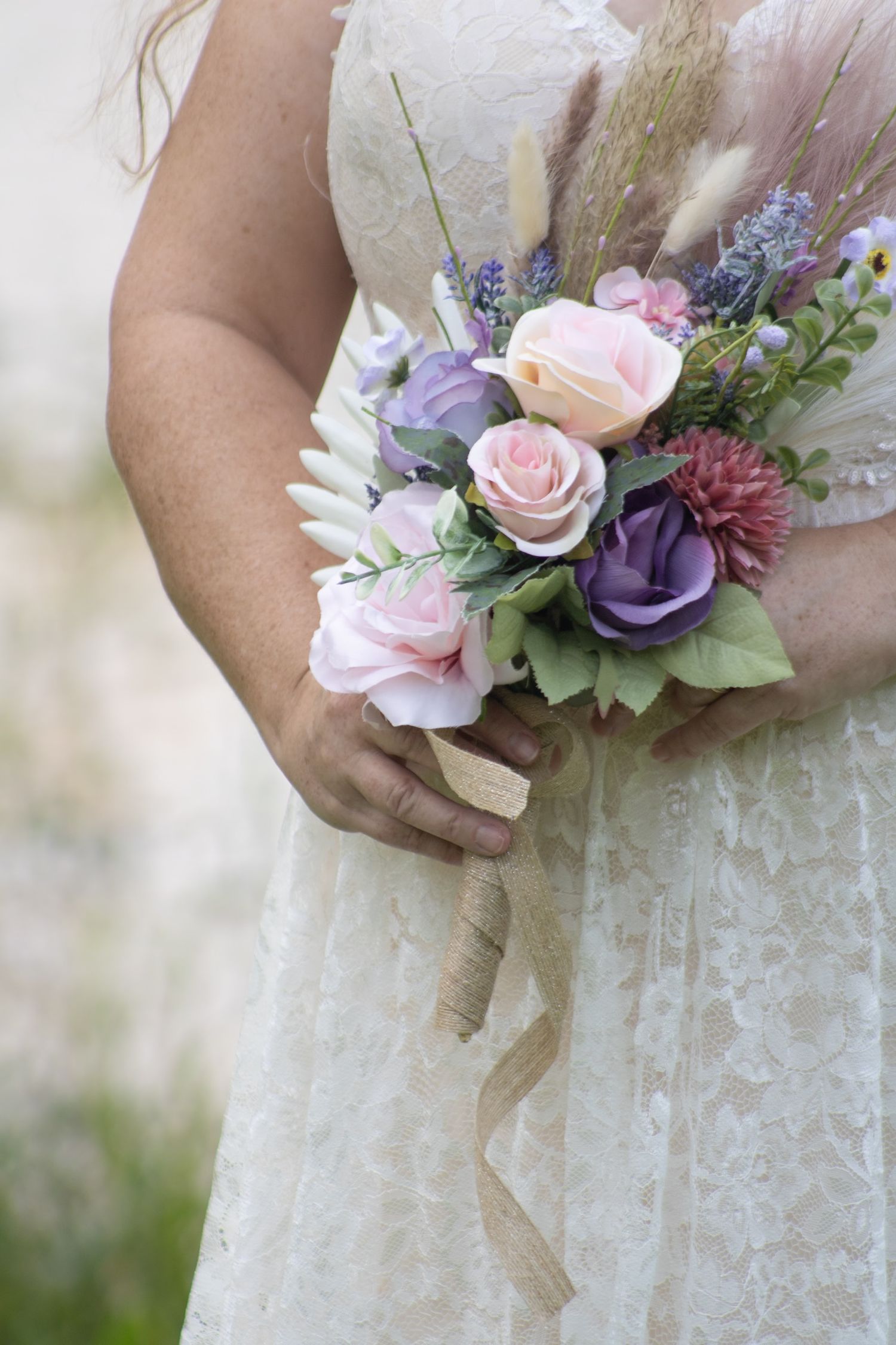 Bride in lace wedding dress holding a bouquet of pink, purple roses and wildflowers with burlap ribbon.