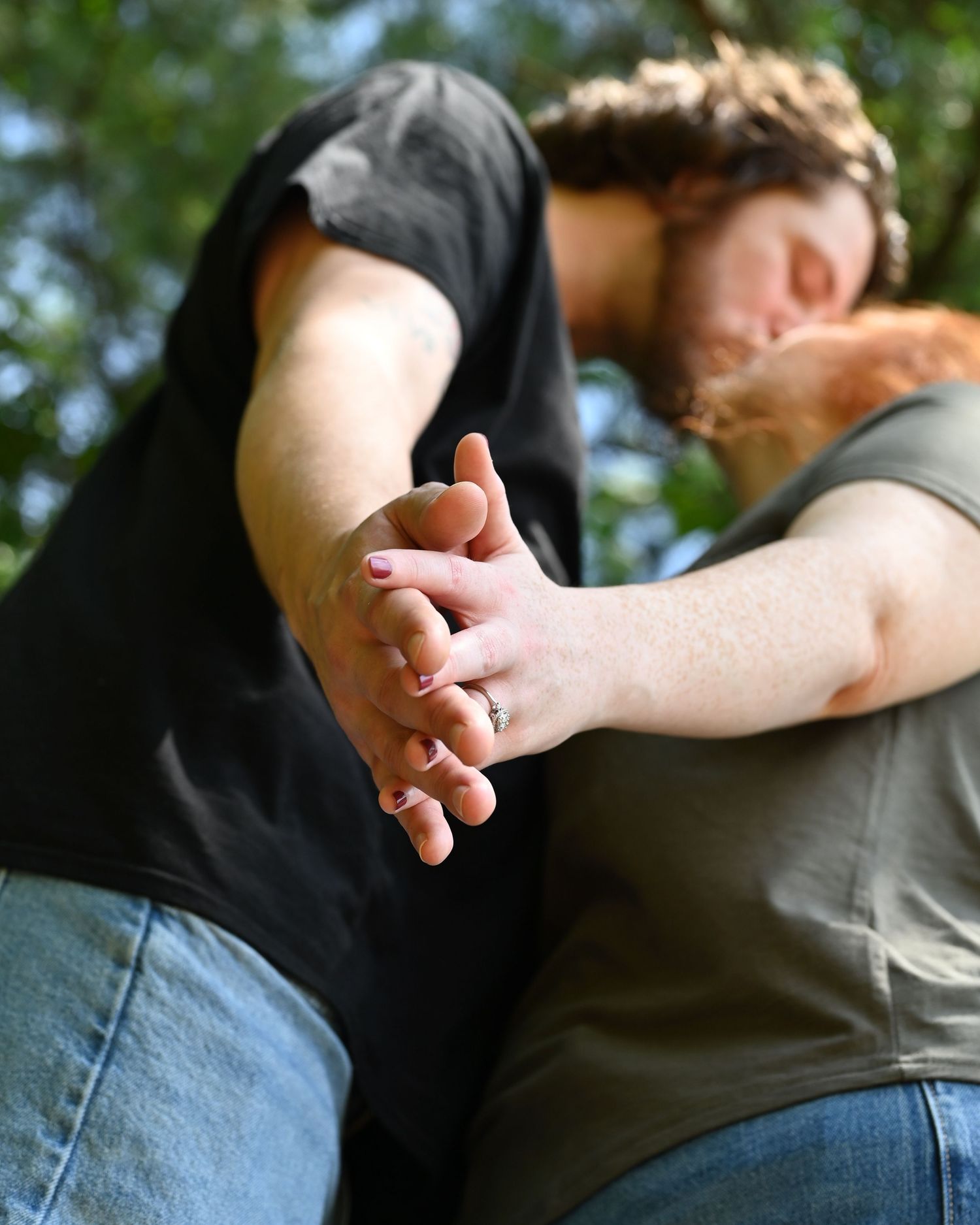 Couple holding hands outdoors, showcasing a silver engagement ring, with trees in the background.