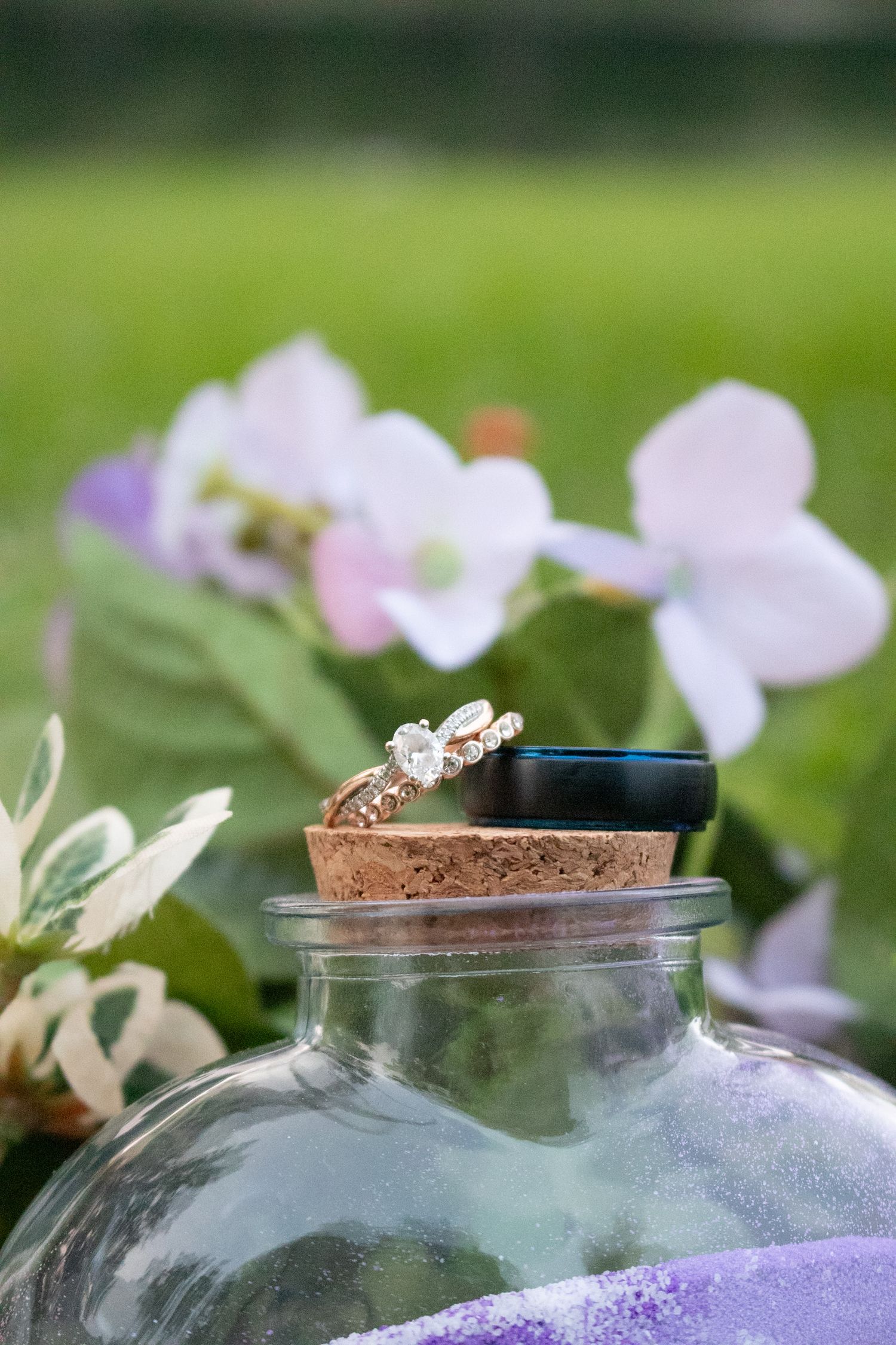 Gold diamond engagement ring and black wedding band resting on a cork bottle top surrounded by purple and white flowers.