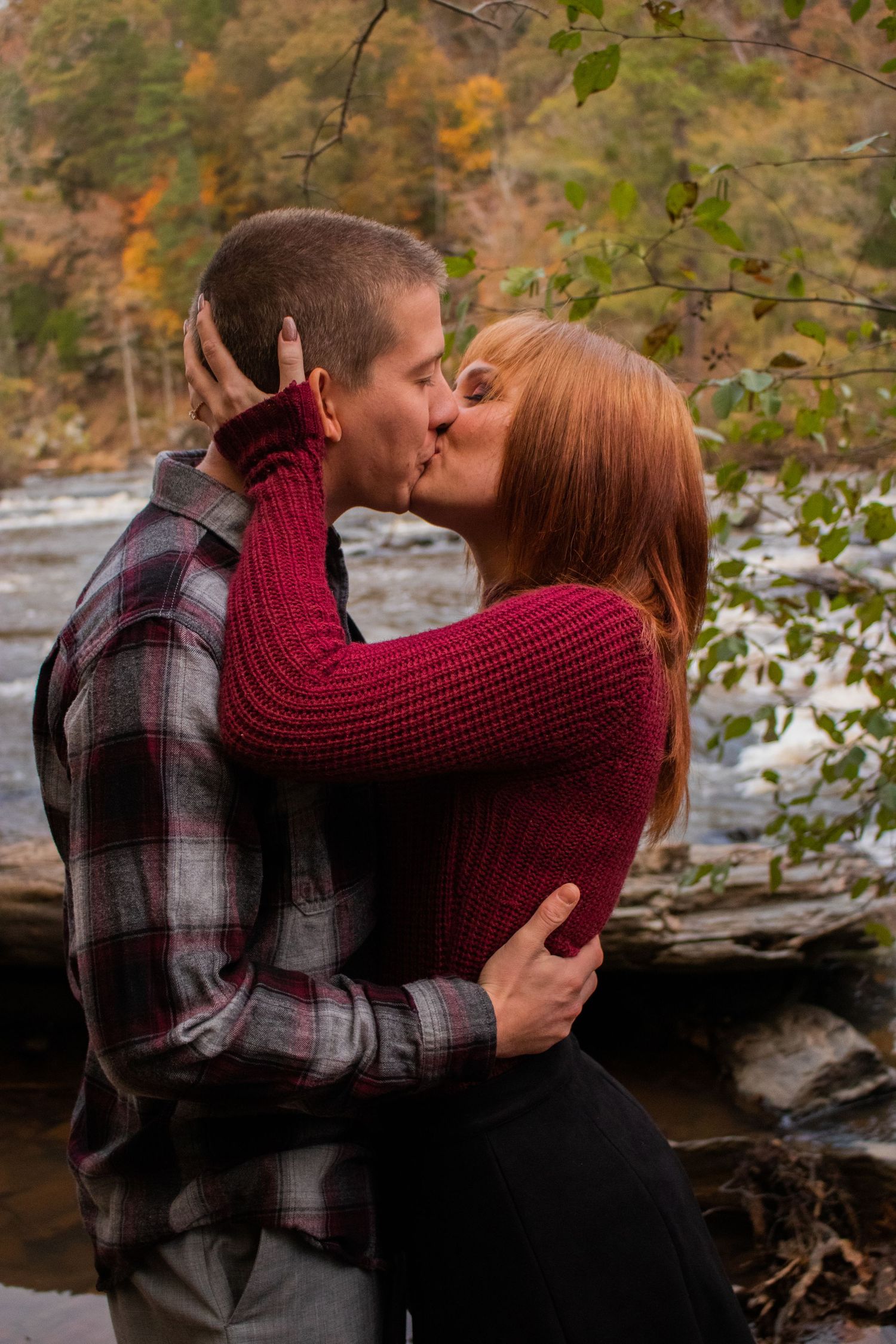 Couple sharing a romantic kiss outdoors by a river in autumn, woman in red sweater, man in plaid shirt.