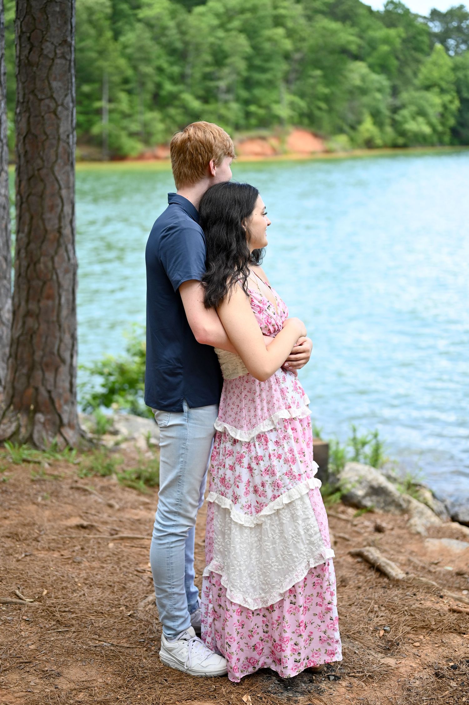 Couple standing together by a serene lake, man in navy shirt embracing woman in tiered floral pink dress.